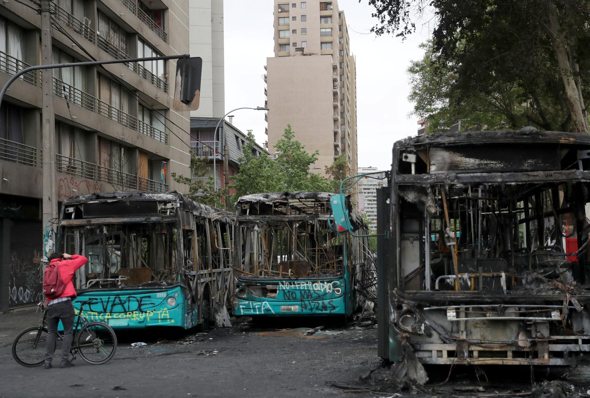 A man is shown with a bicycle standing in front of three totally burned out buses in a street.