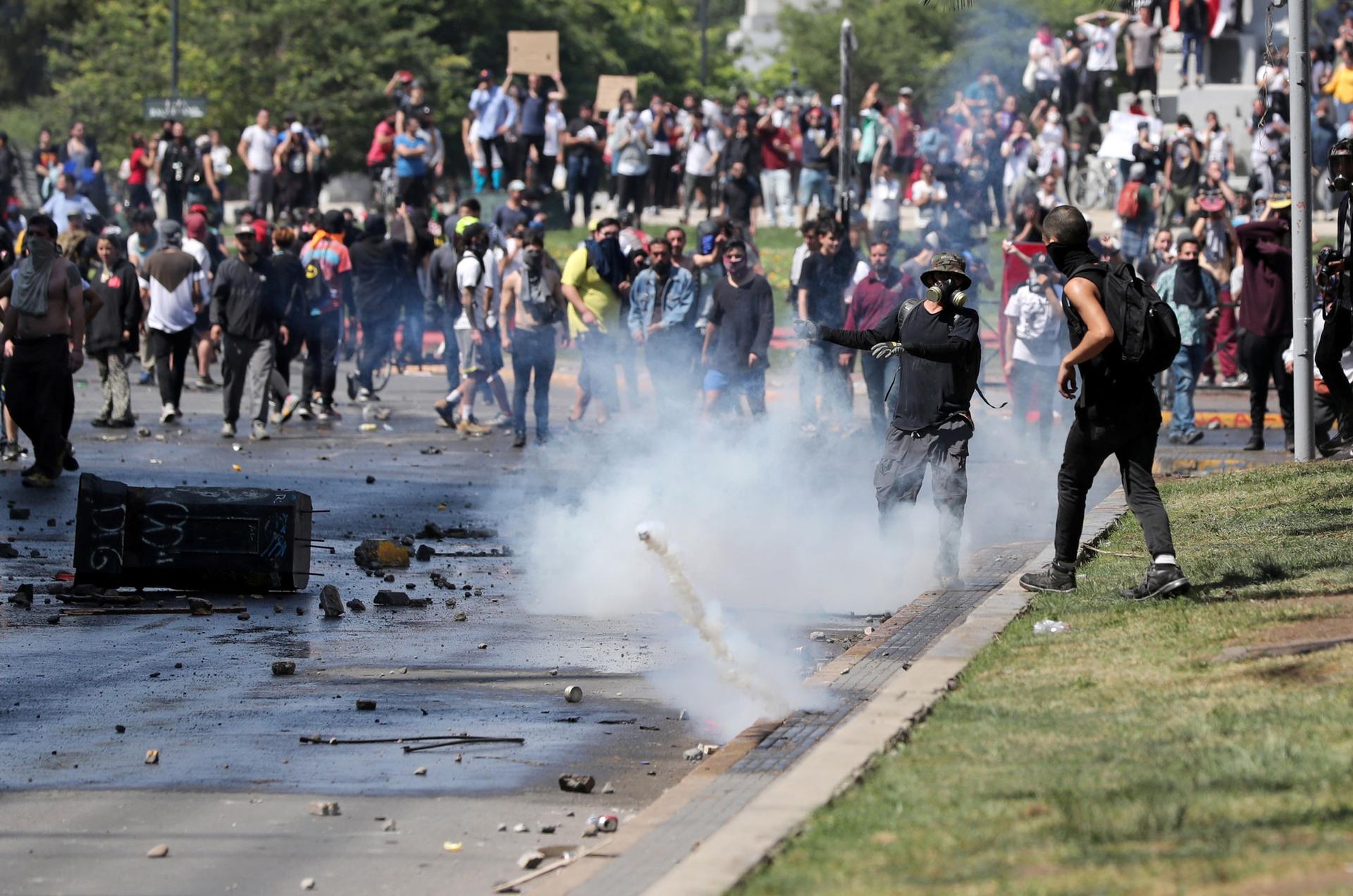 A large group of protesters are shown in the street with a smoking tear gas canister bouncing through the middle of the group.