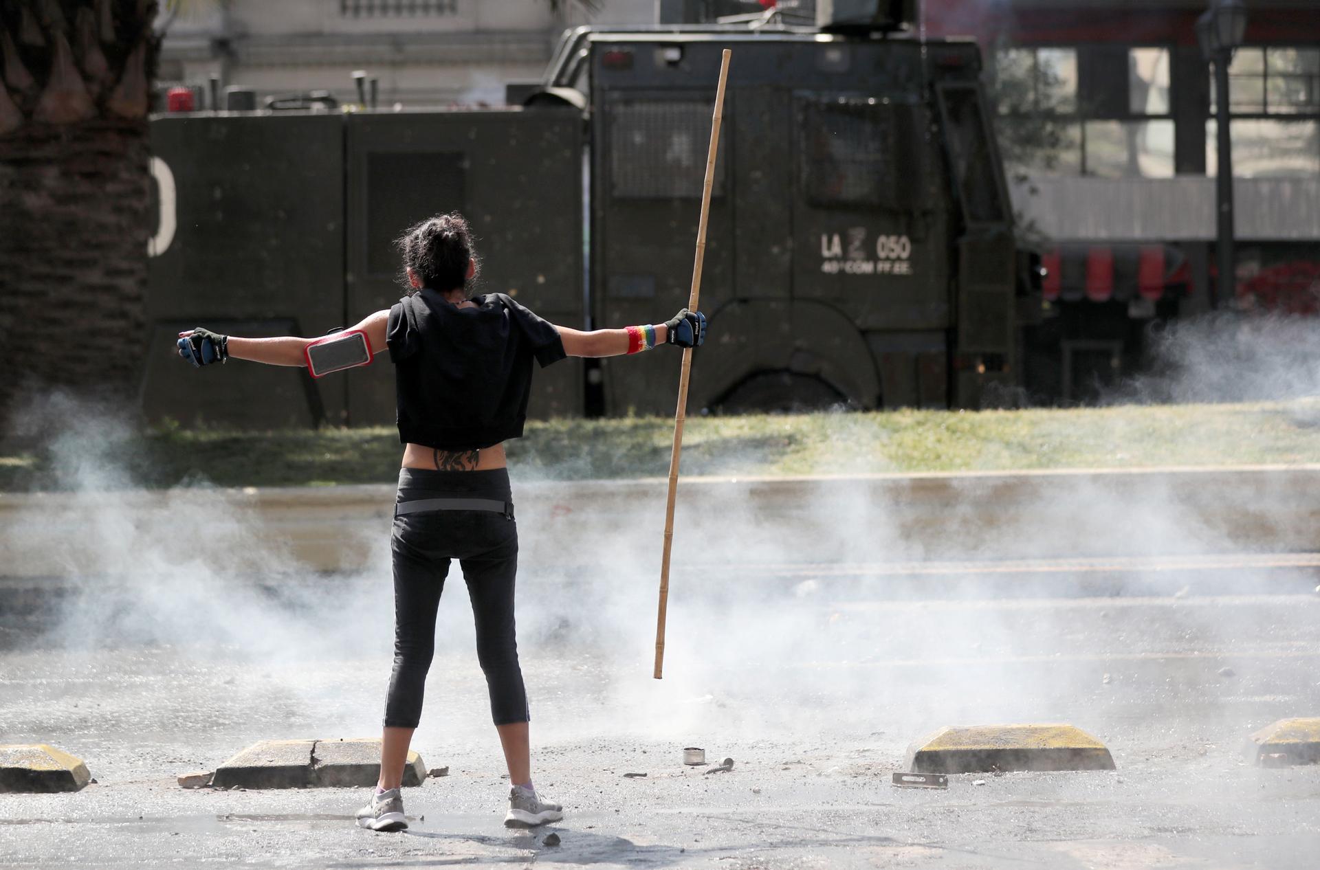 A woman is shown stands with her arms out, holding a stick, with smoke around her and a military truck in the distance.