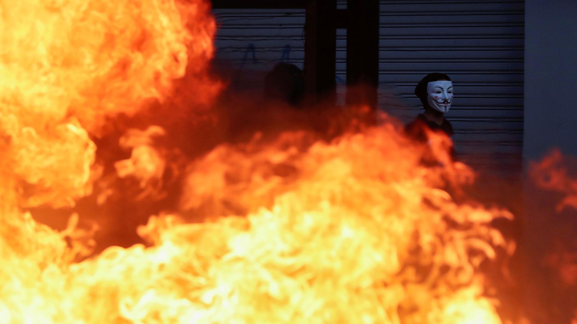 A demonstrator wearing a Guy Fawkes mask is shown walking past a large fire.