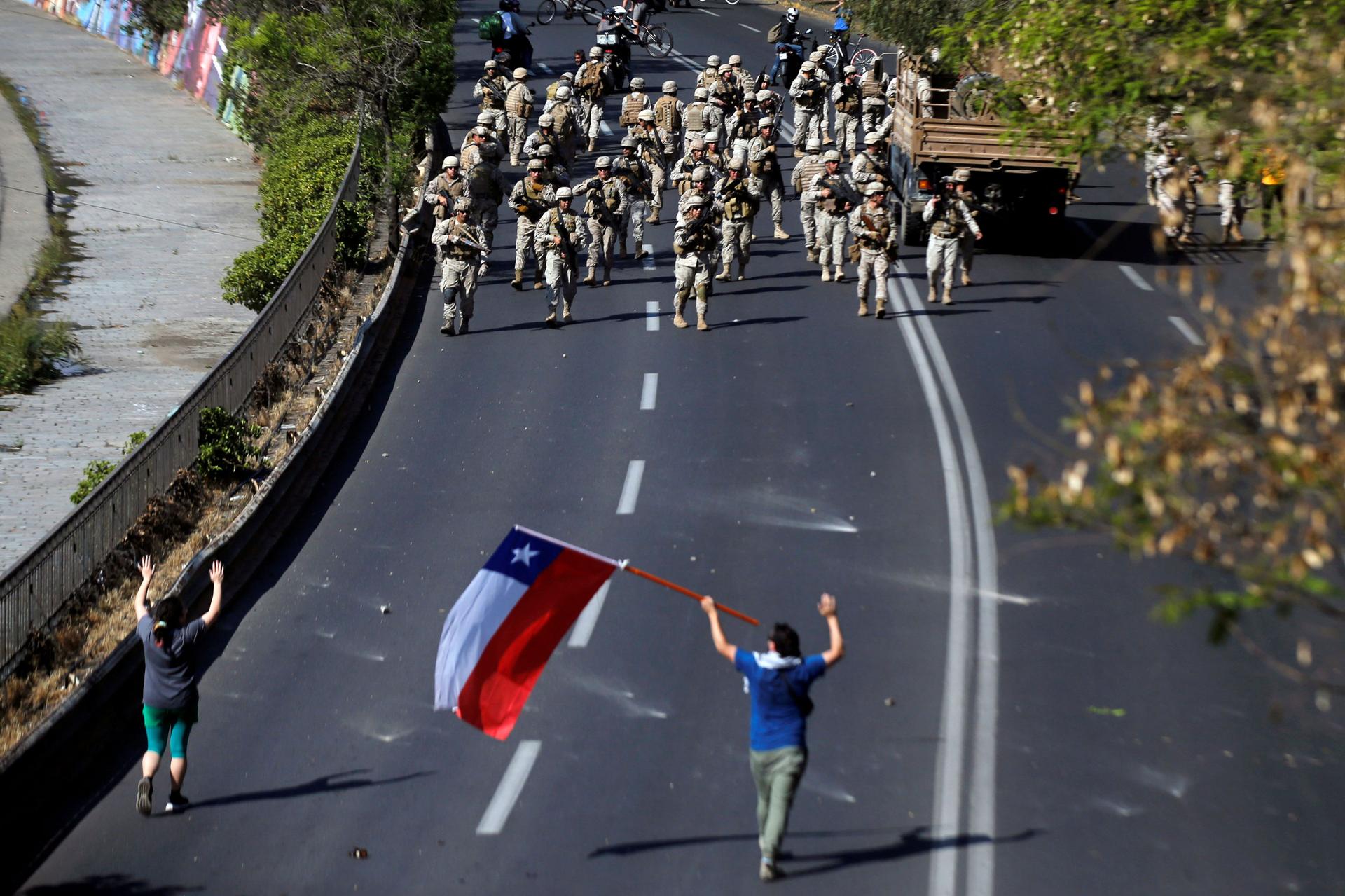 Dozens of Chilean soldiers are shown marching down a road toward two protesters, one carrying a Chilean national flag.