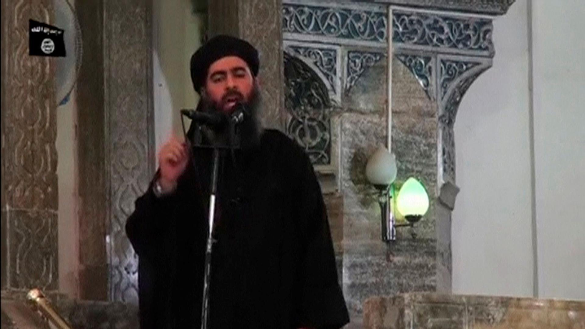 Abu Bakr al-Baghdadi is shown in a black shawl and turbine speaking into a microphone.