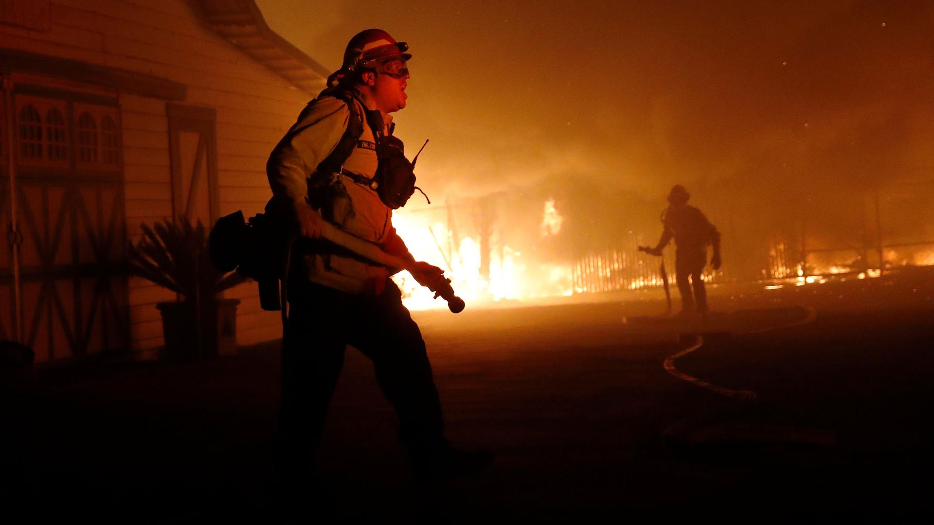 A firefighter is shown in the nearground with a barn and other buildings are on fire in the background.