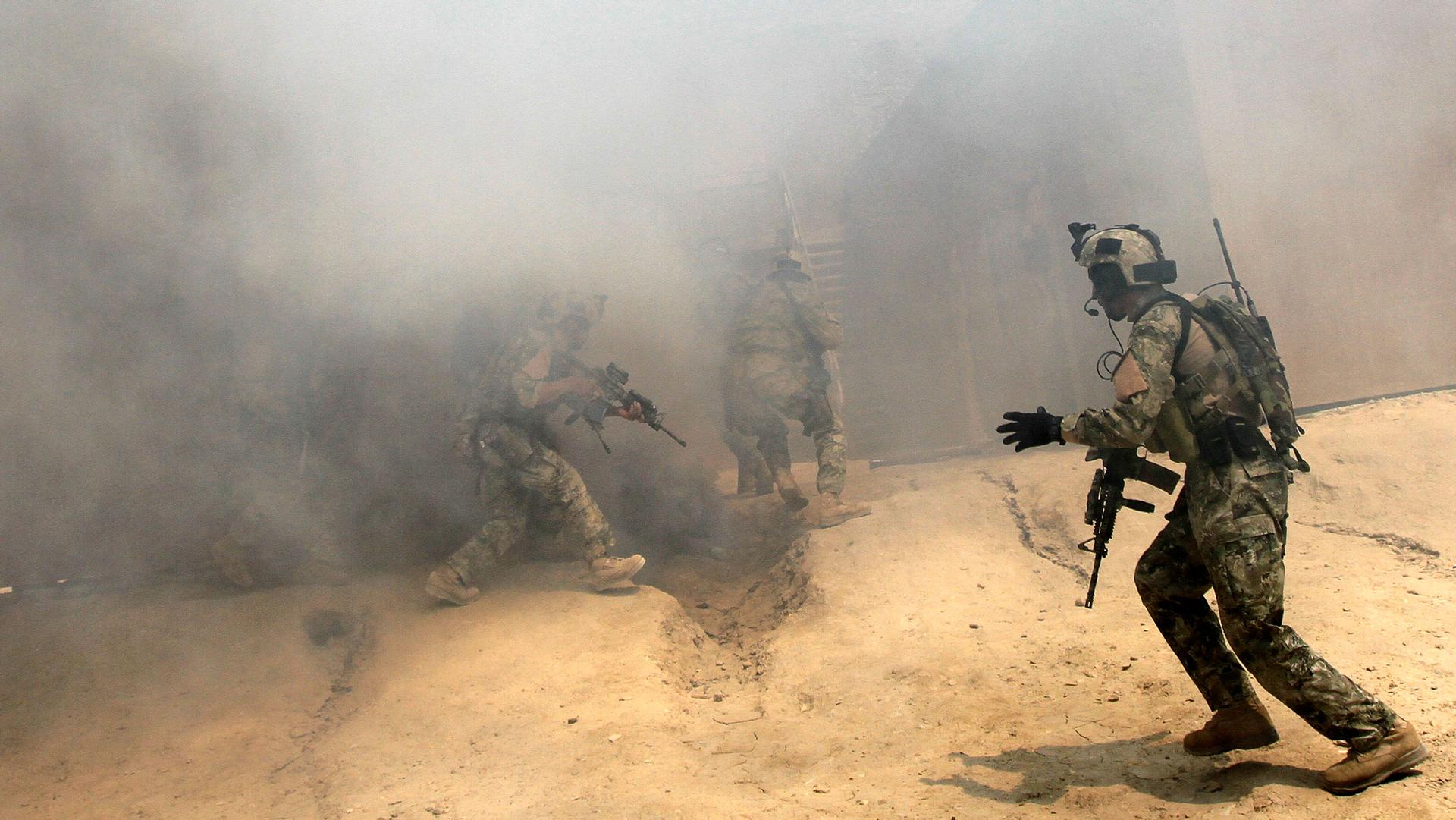 Men in military uniforms hold weapons and are walking into smoke inside building.