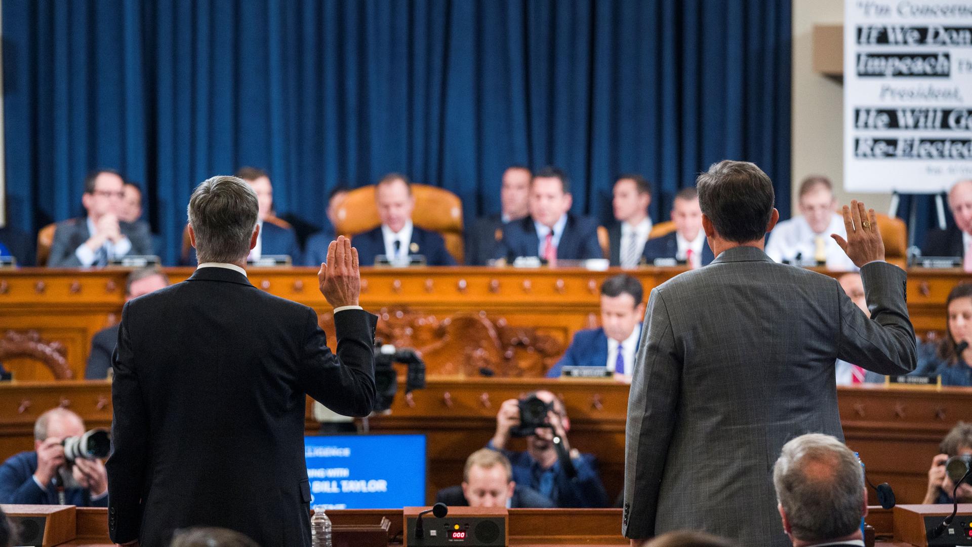Two men stand with their backs to the camera, their right hands held in the air for an oath. Ahead of them is a row of lawmakers seated behind a large desk.