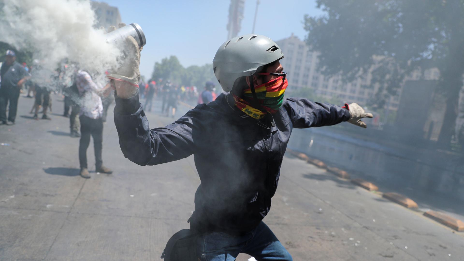 A protester with a bandana covering their face and wearing a helmet, cocks an arm back to throw a smoking canister of tear gas back at police.