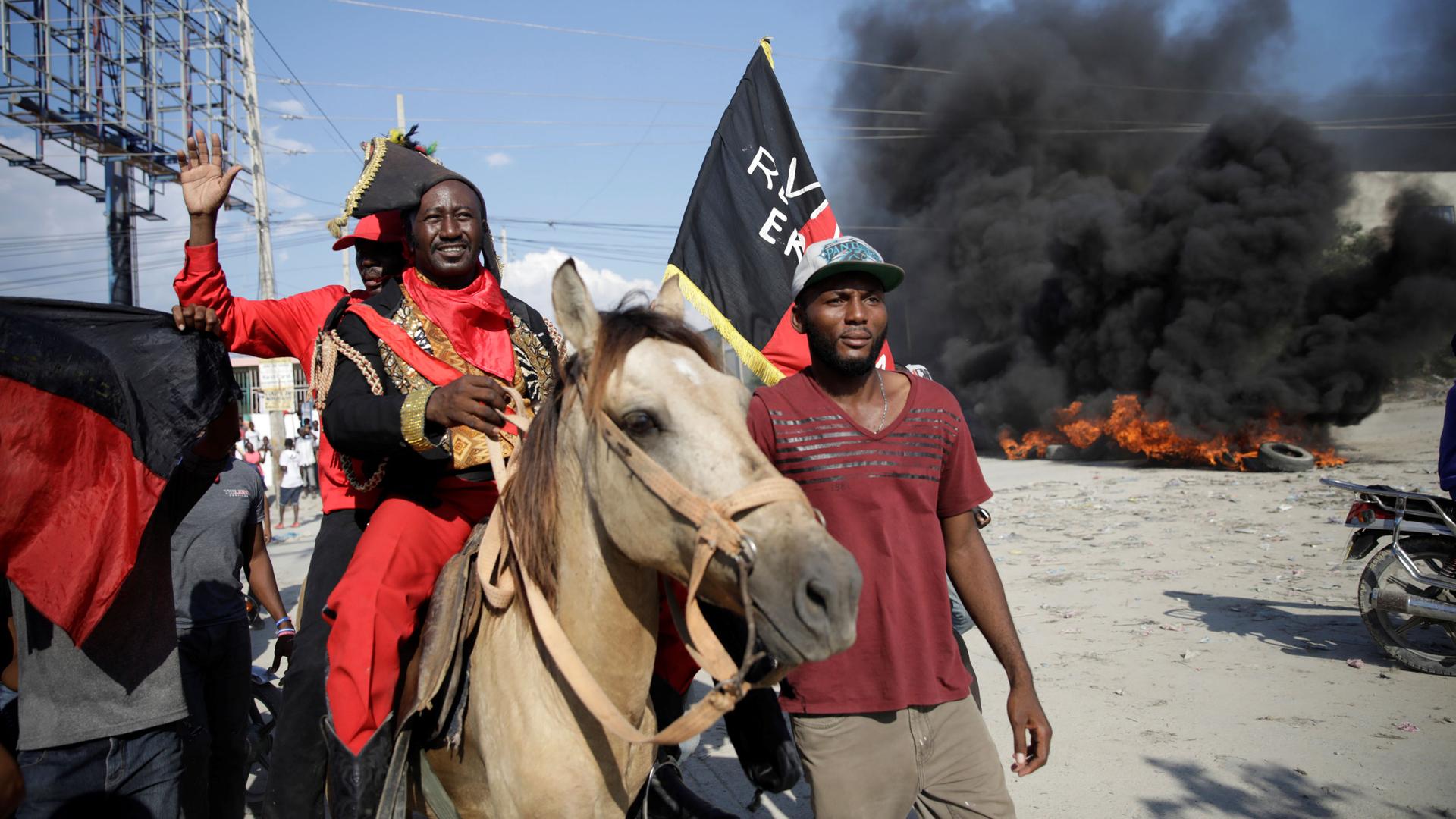 A man on a horse wears a black and red colonial-era costume. Behind him is smoke from a fire. 