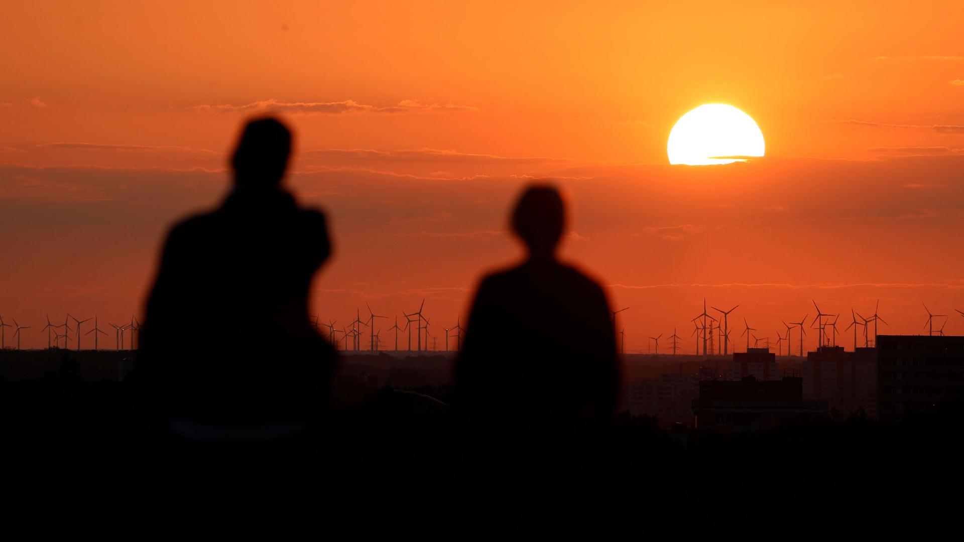 A bright orange sunset is shown in the distance with several wind turbines in the distance and two people in shadow in the nearground.