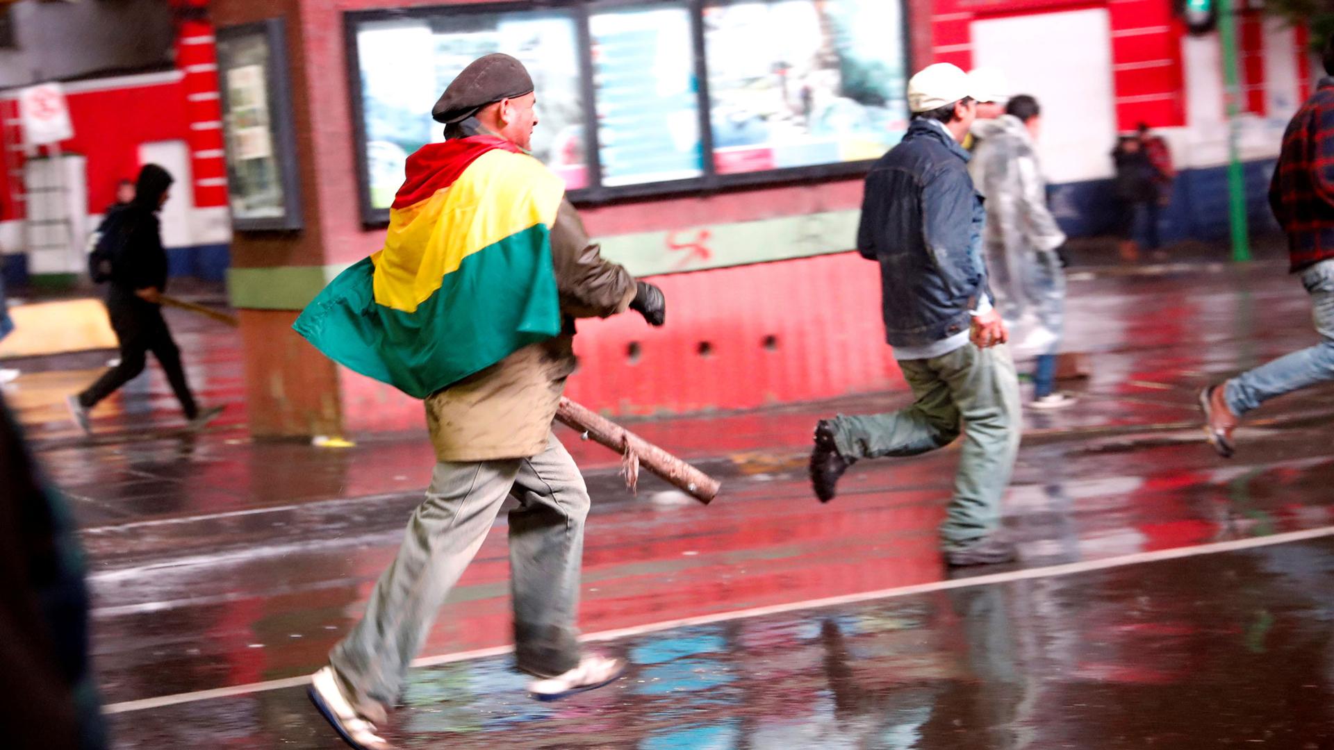 Several people are shown running in the street with one man draped in a Bolivian flag.