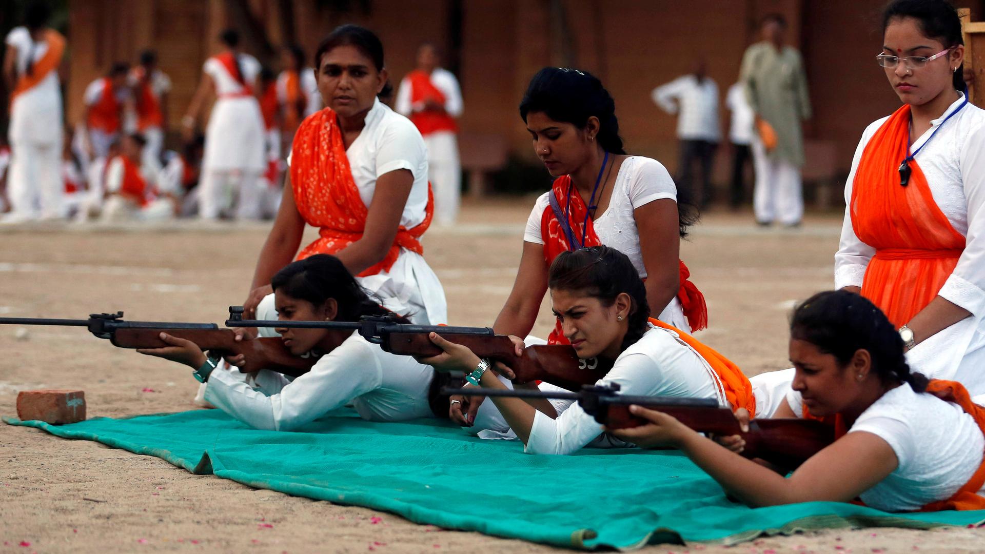 Women lie on a green cloth and practice aiming their guns while wearing red sashes. 