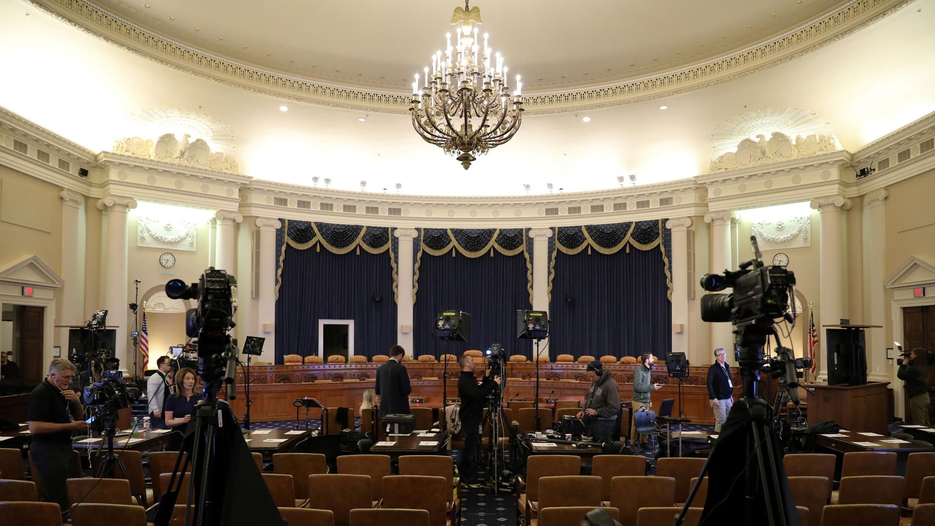 A House committee room is shown with a chandelier hanging from the ceiling and two television cameras placed among the seats.