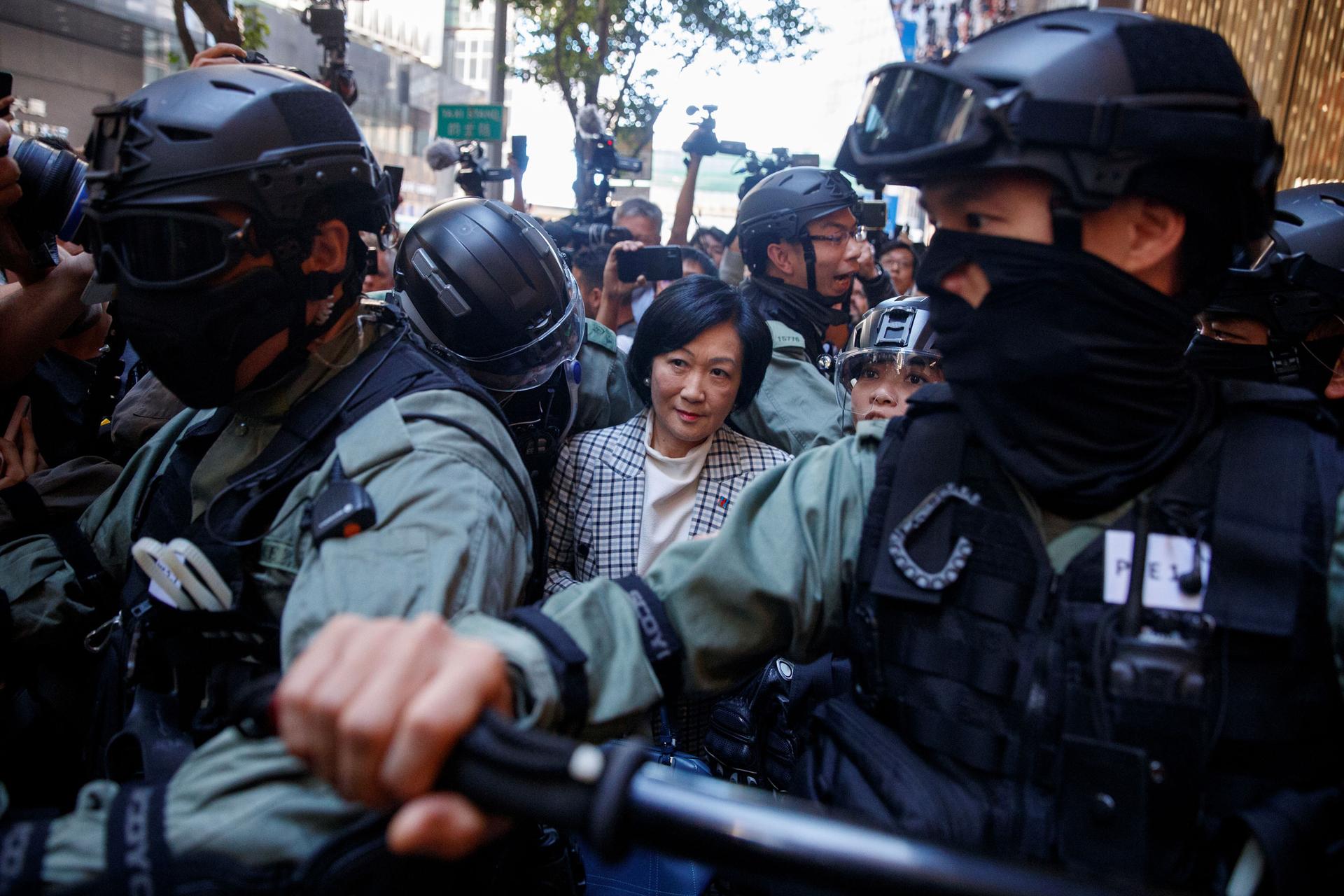 A woman is shown wearing a checkered suit coat and surrounded by an armed police escort.