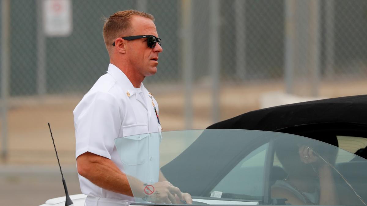 A man in a white navy uniform and black sunglasses walks into a car