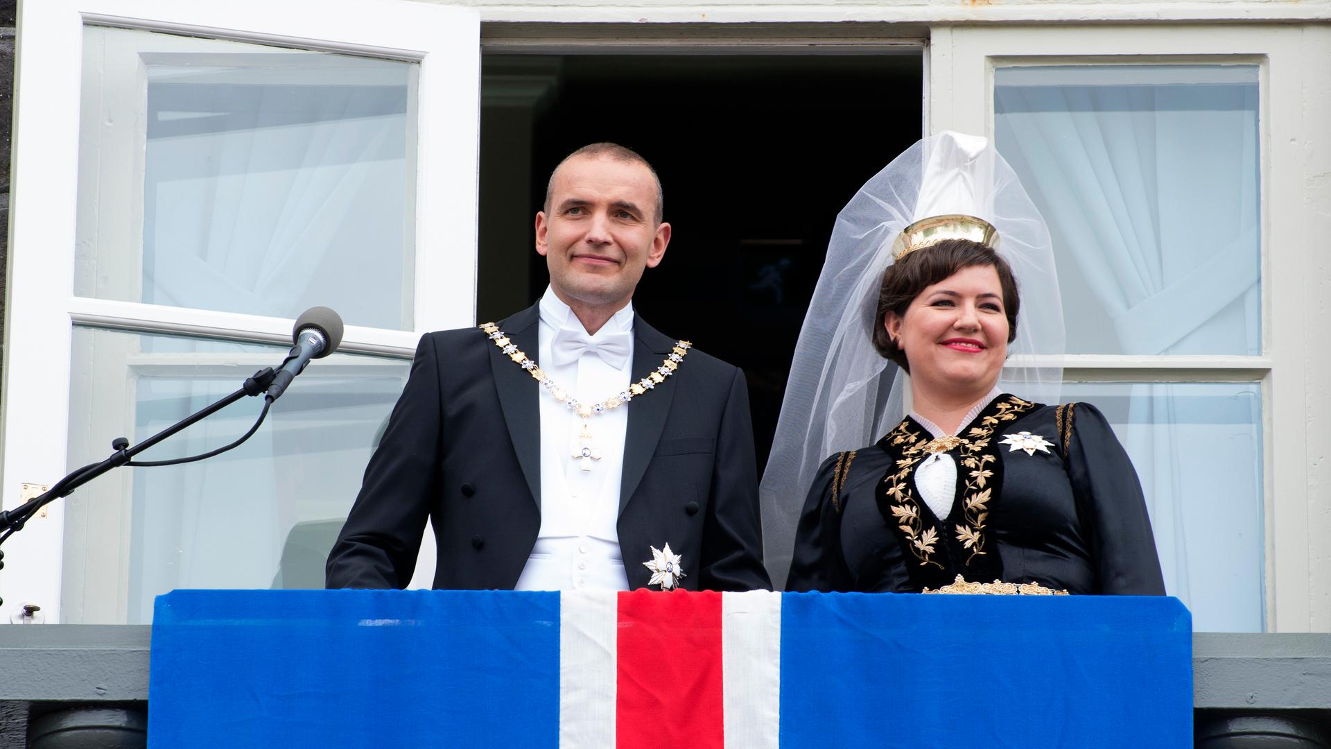 Man and woman wearing crown stand on balcony with blue, white and red flag in front of them