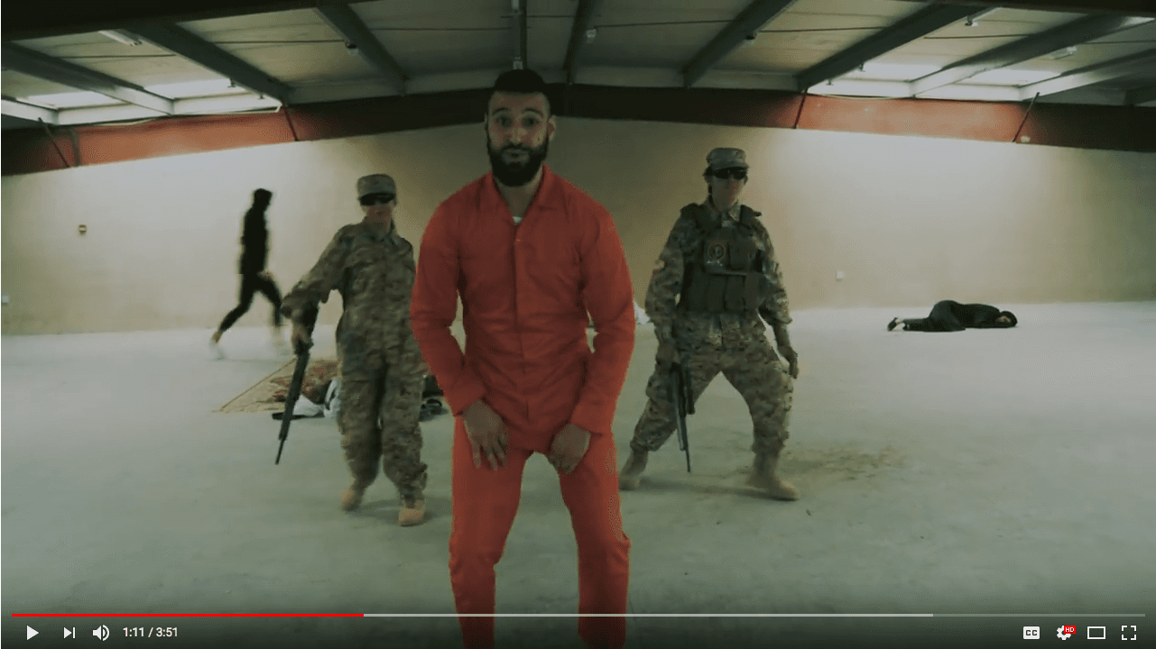 Man in orang jumpsuit dancing in front of two people in army fatigues