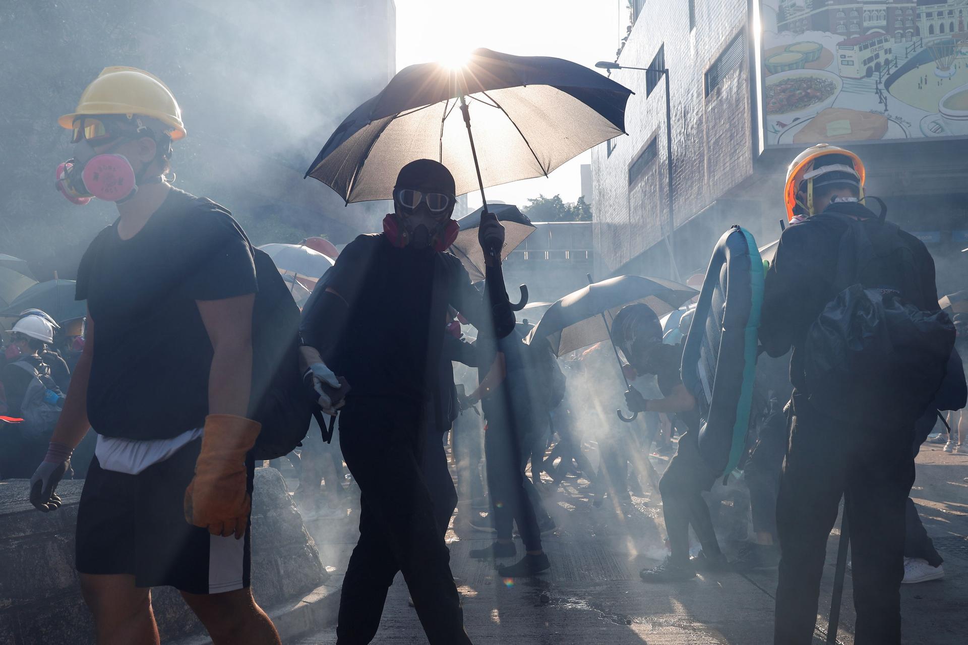 Several protesters are shown wearing gas masks and one holding an umbrella with the sun glaring above it.