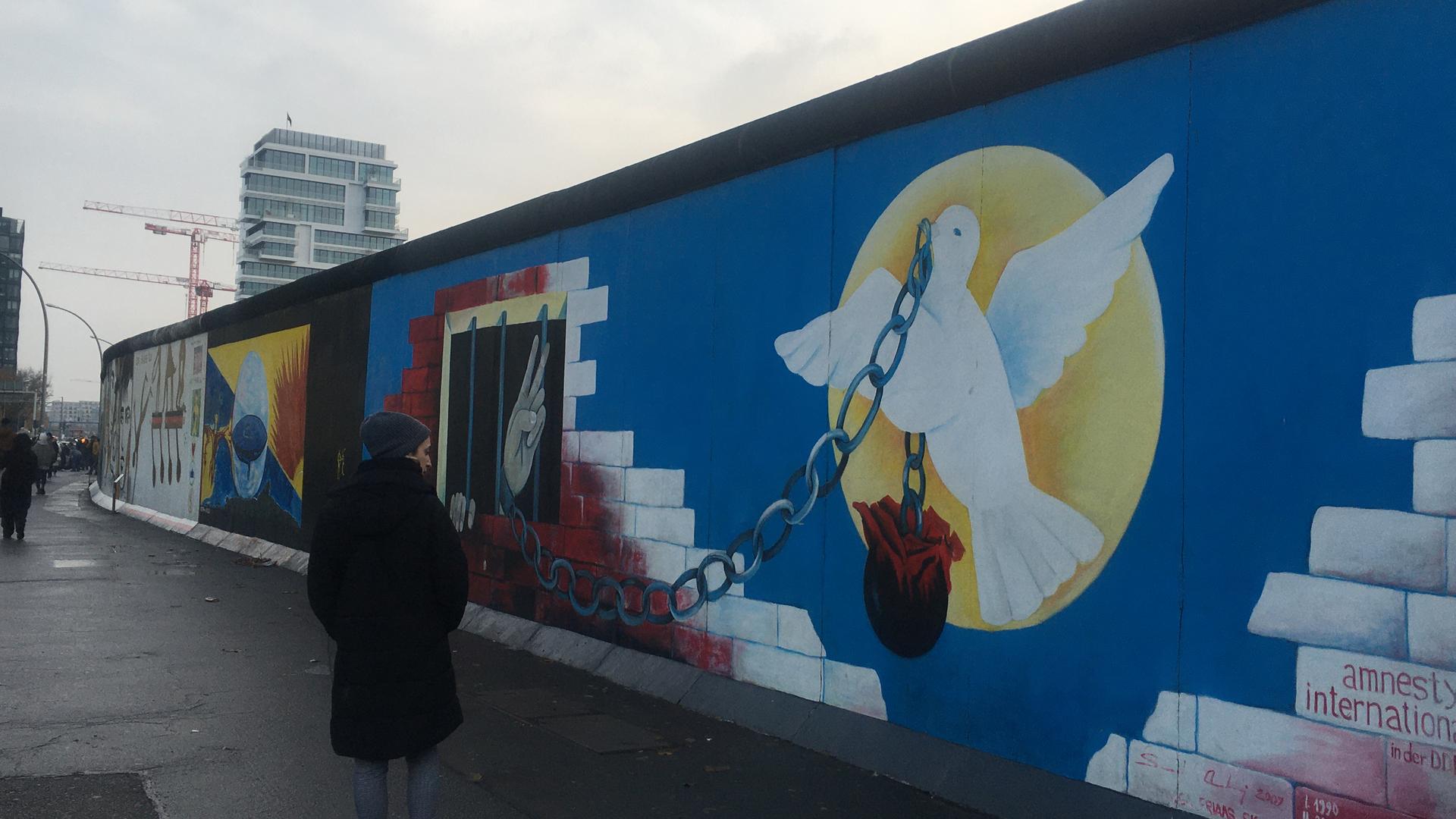 A mural of a white bird, chain and rose painted on the Berlin wall and a person walking by it