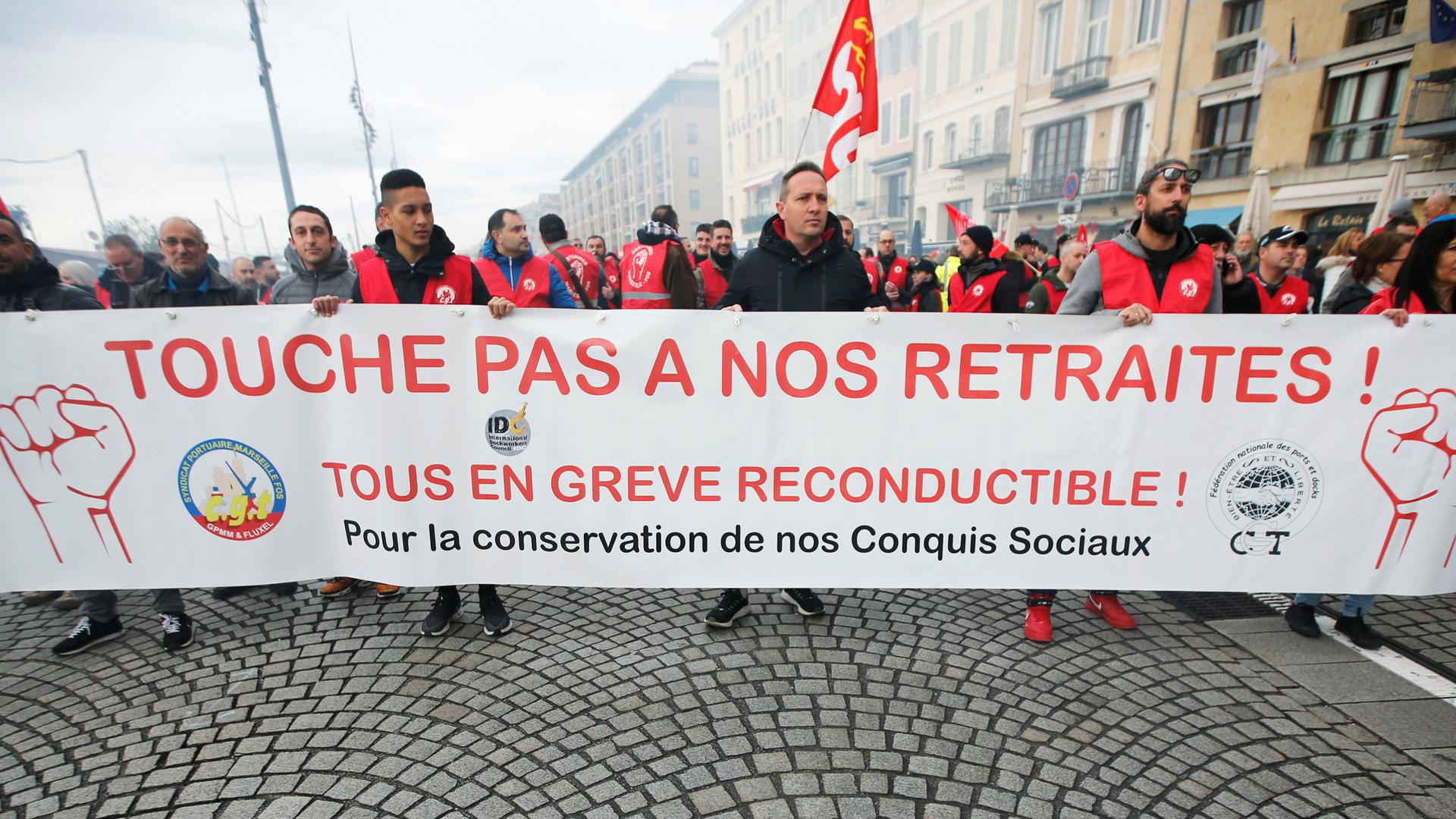 A large group of protesters are shown in the street holding a banner during a demonstration in Marseille. The banner reads, 