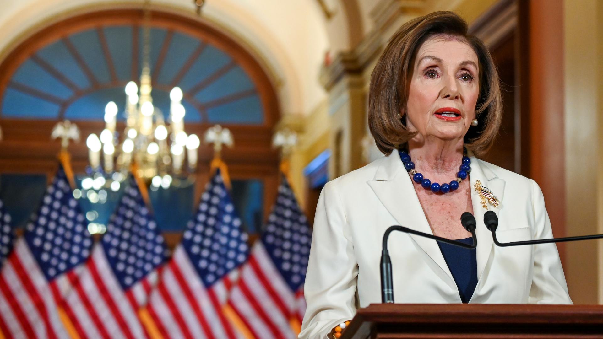 Nancy Pelosi in a white jacket stands at a podium in front of American flags. 