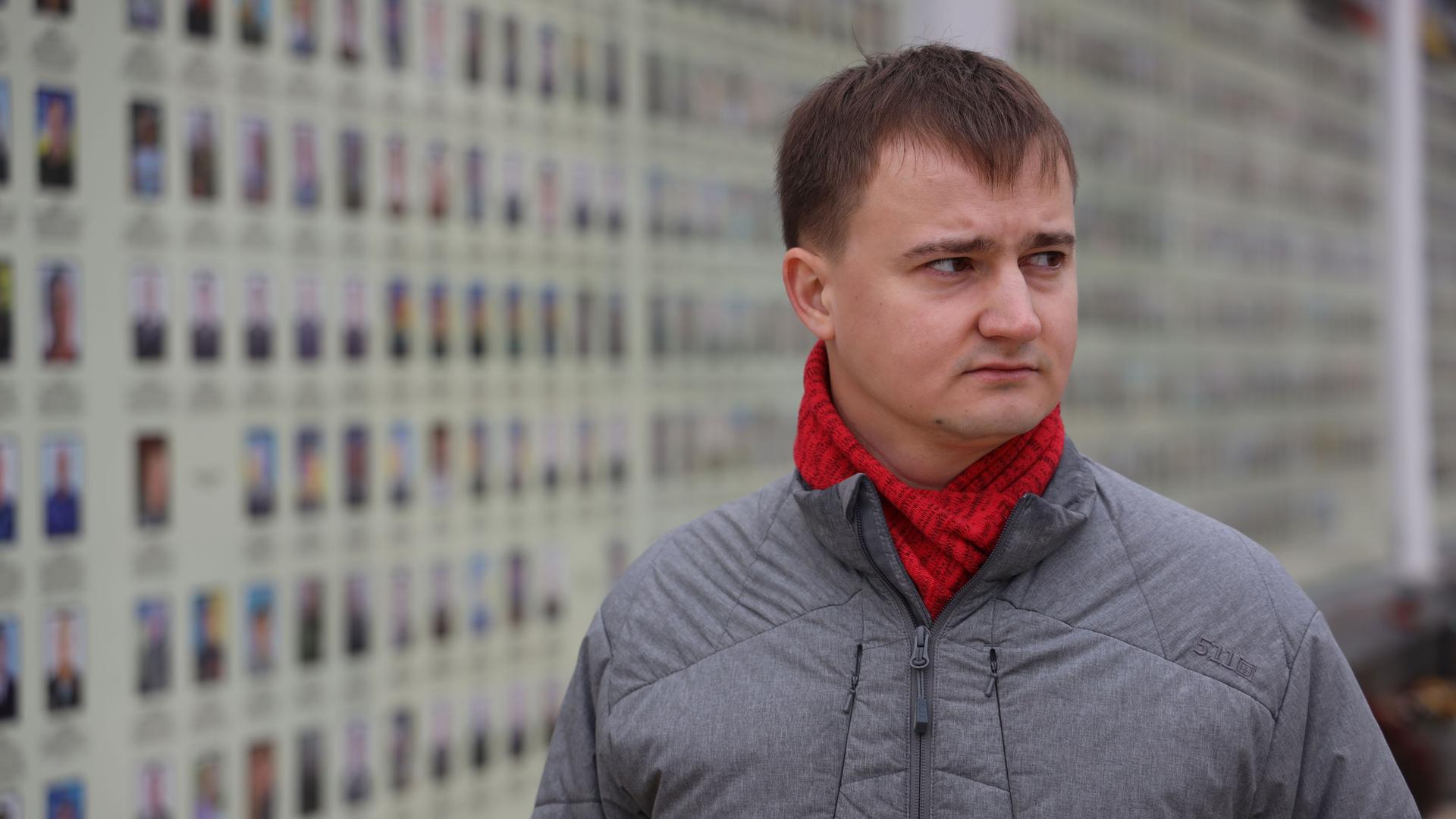 A man stands somberly in front of a wall of small portraits of soldiers