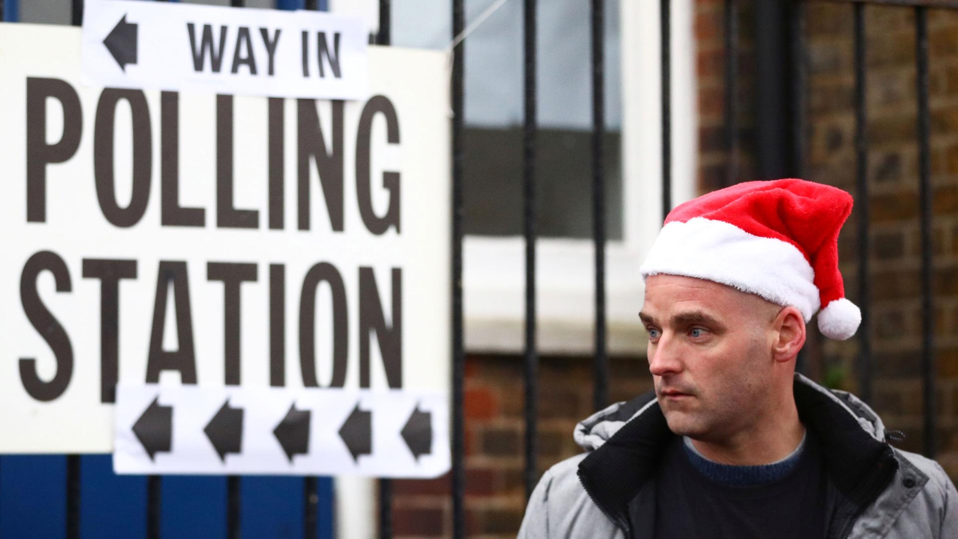 A man is shown wearing a red and white Santa Claus hat and standing next to a white sign with the words 'polling station' printed on it.