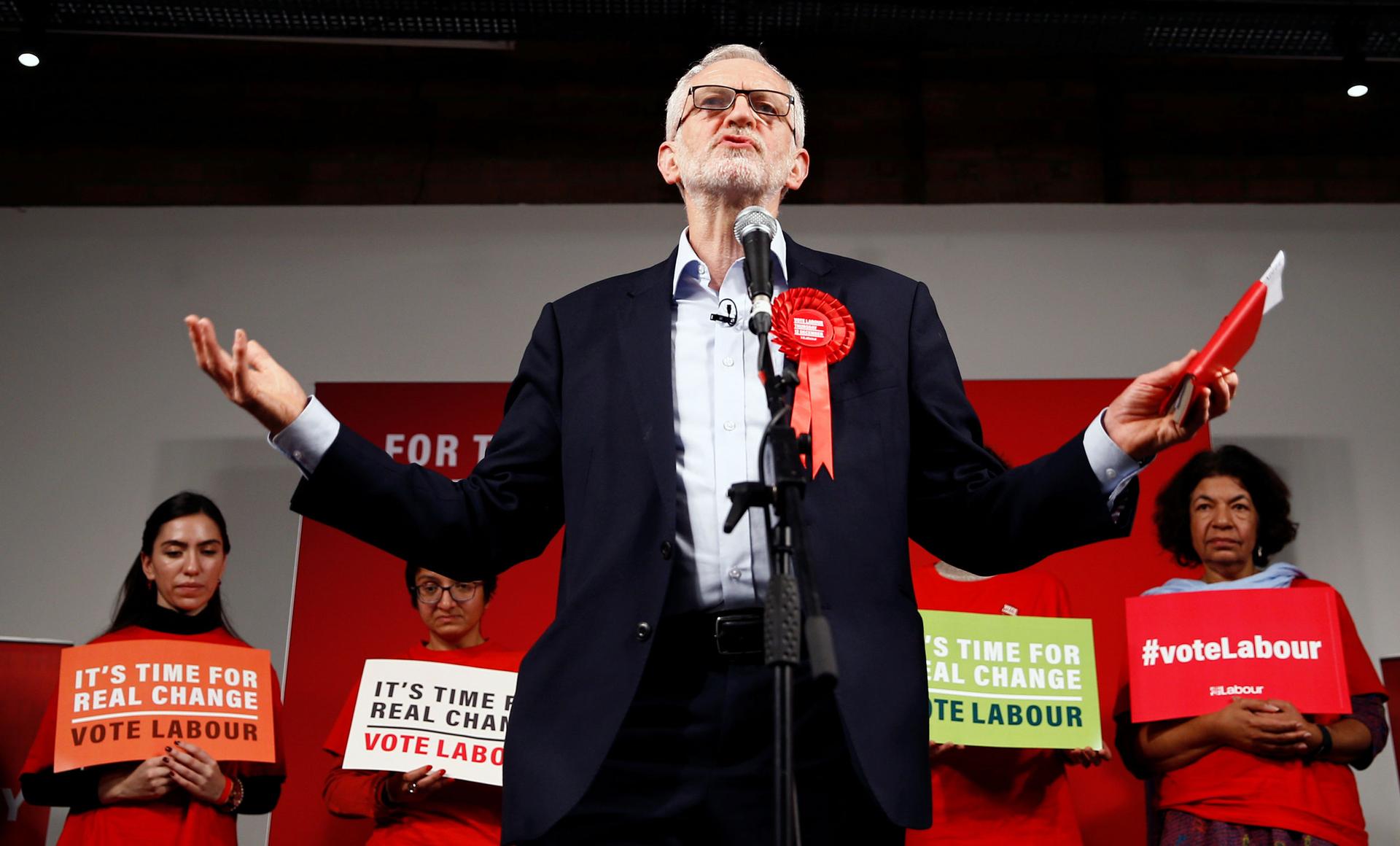 Britain's opposition Labour Party leader Jeremy Corbyn is shown standing a microphone wearing a large red ribbon and holding a red book.