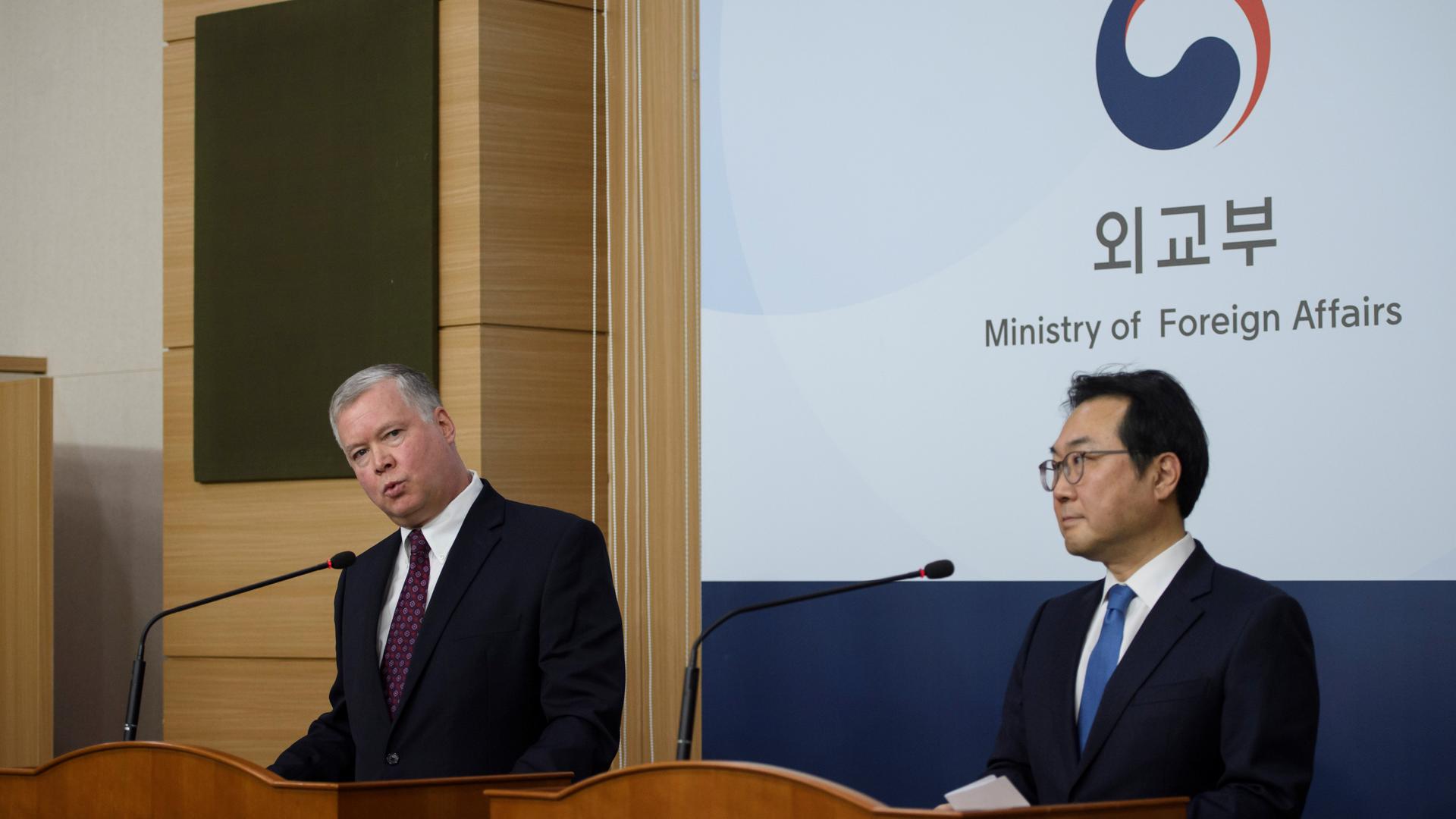American man wearing dark blue suit speaks at podium standing next to Korean man wearing dark blue suit. 