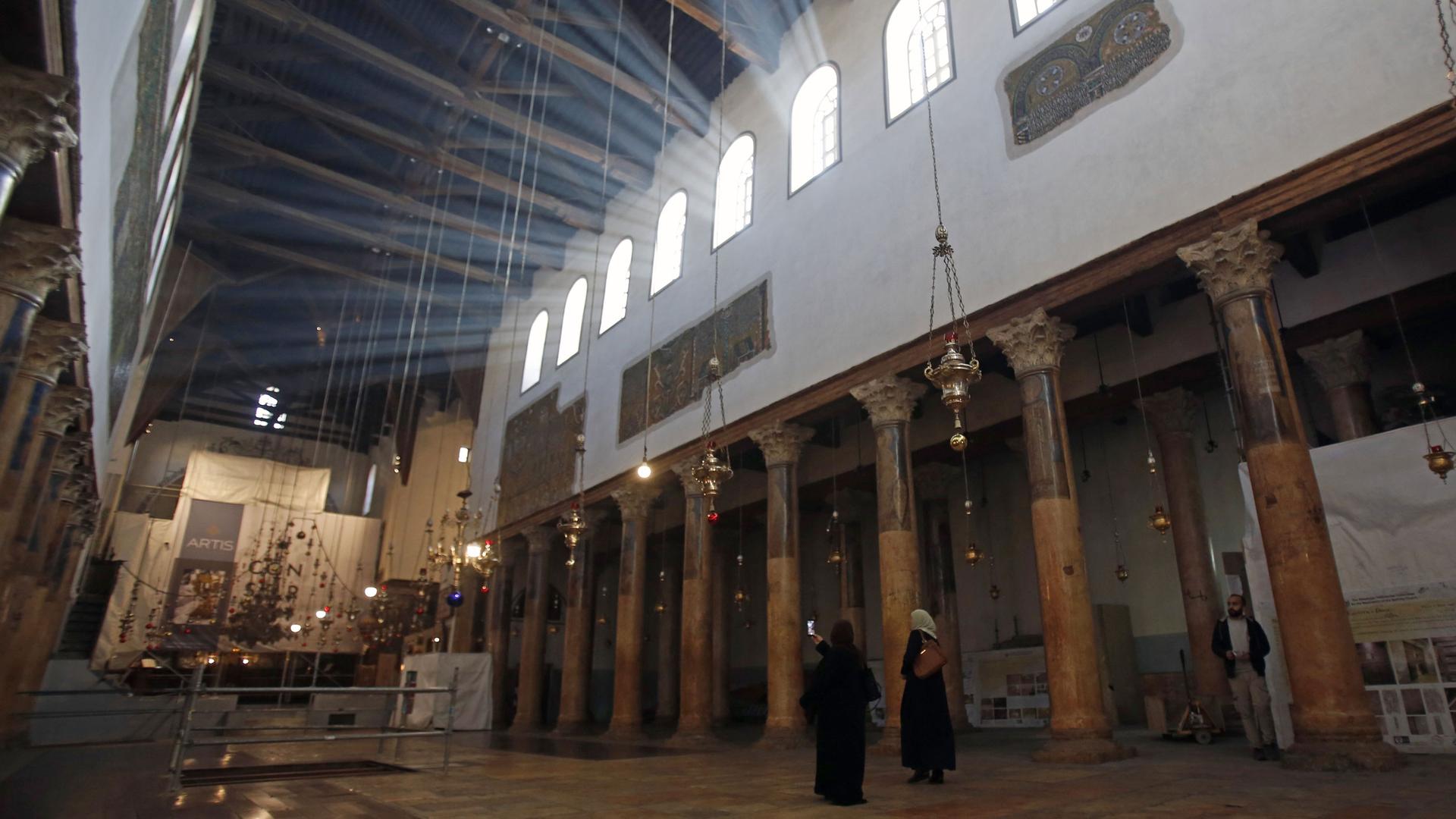 Two visitors are shown standing a one taking a cell phone photo of the Church of the Nativity with bright white light shining from the windows above.