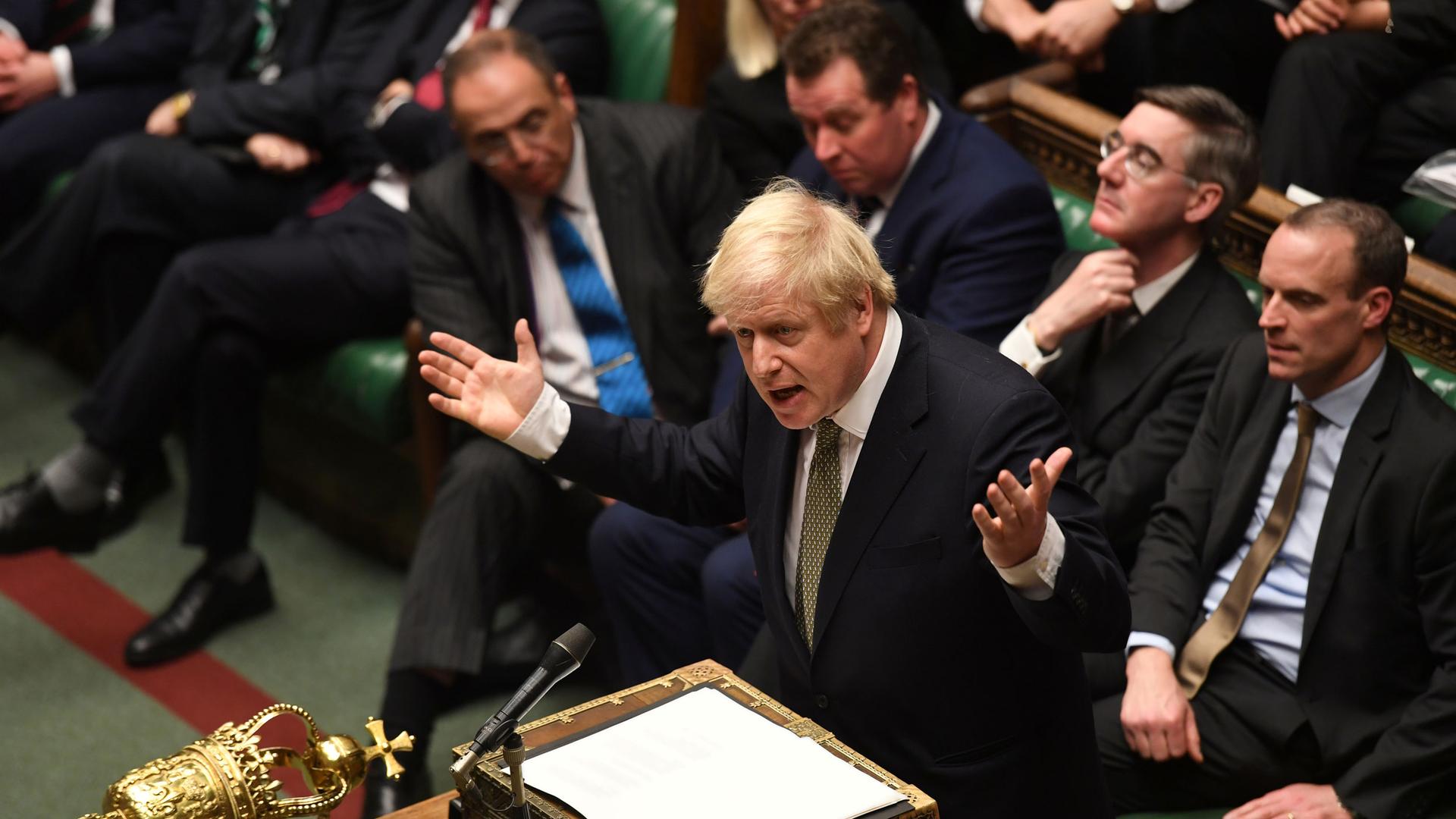 Britain's Prime Minister Boris Johnson is shown standing at a lecturn with a microphone and several parliamentary members sitting behind him.