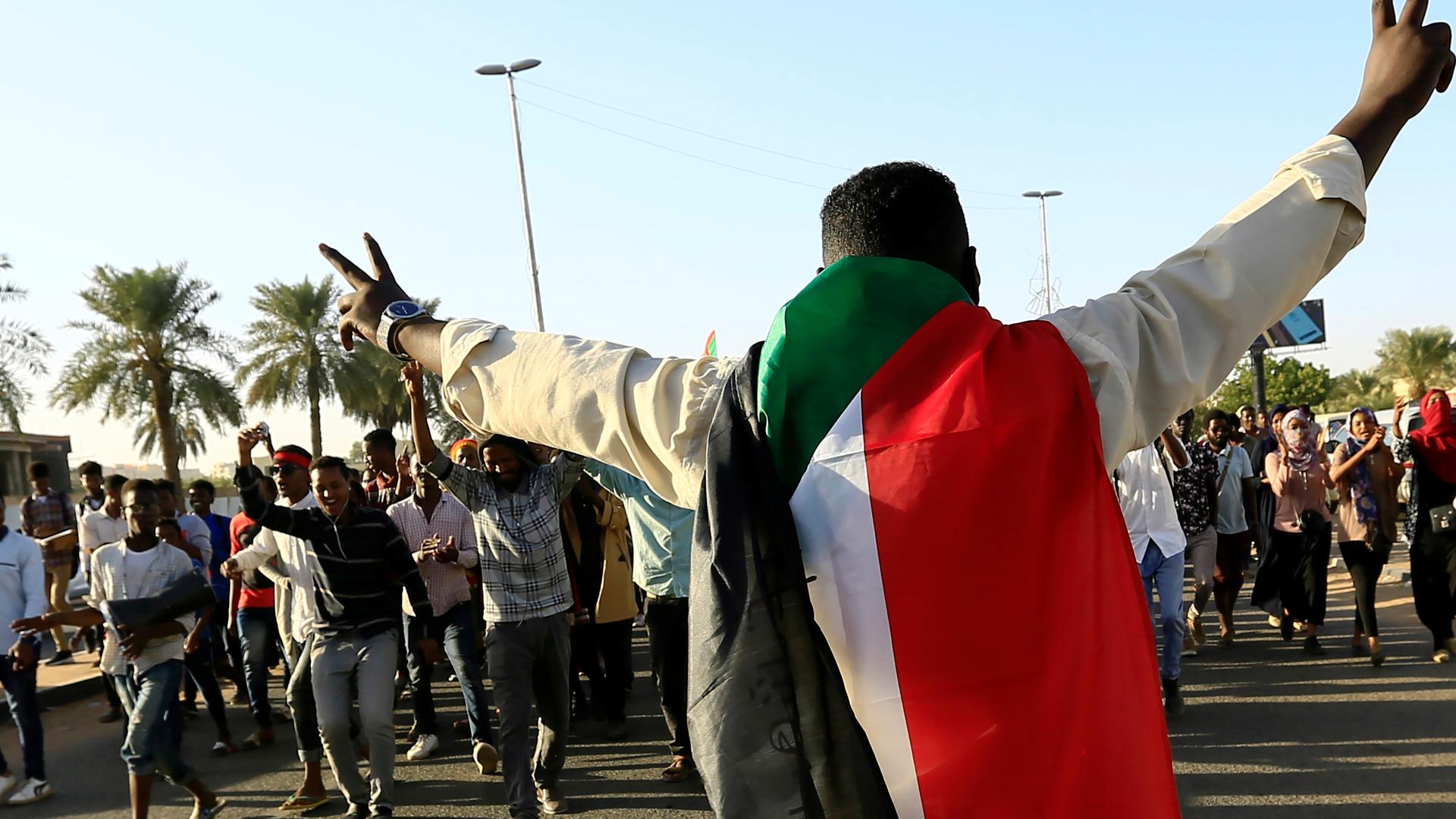A protester holds peace sign and wears Sudanese flag across his shoulders