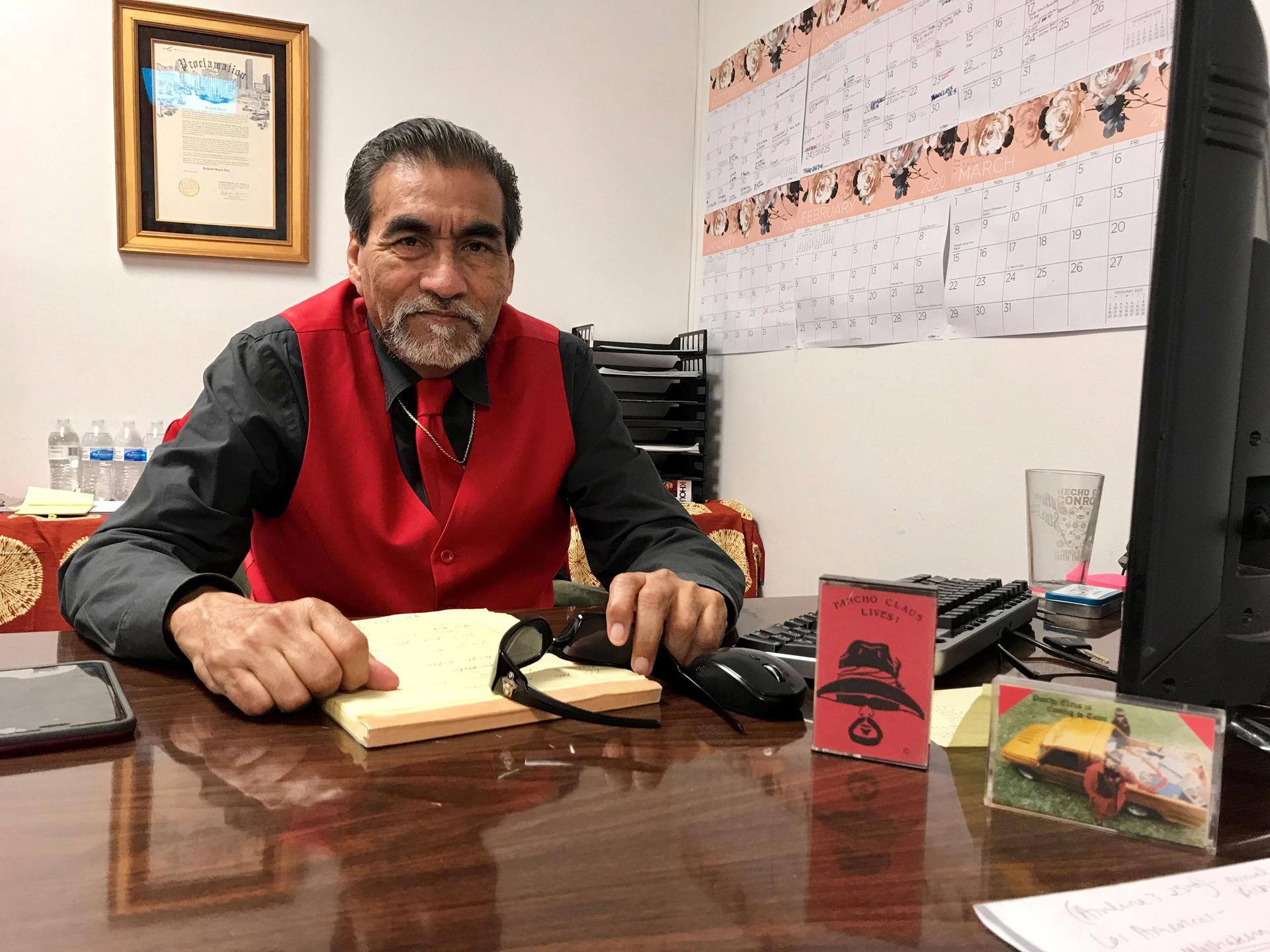 Richard Reyes is shown wearing a red vest and tie while sitting at his desk