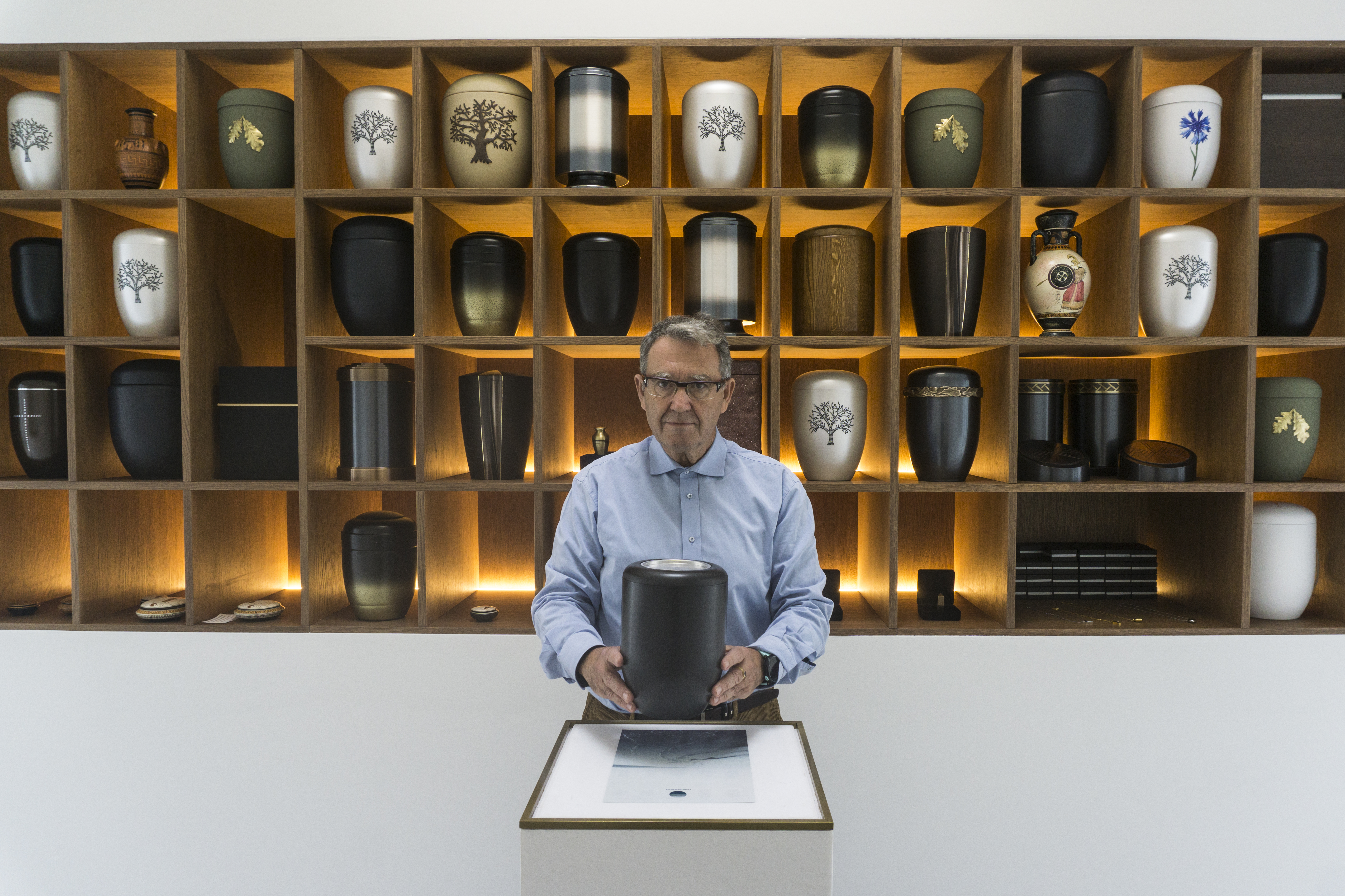 A man stands at a counter holding an urn with many urn designs behind him