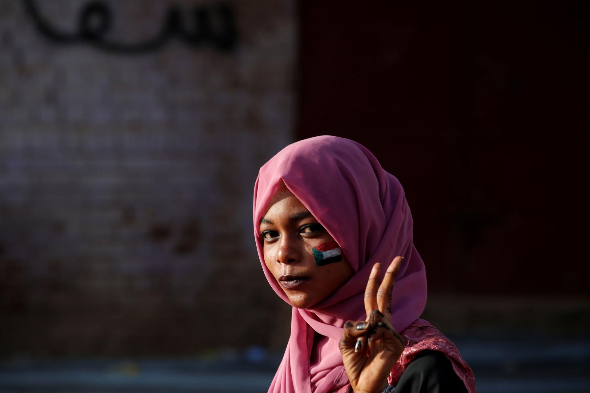 A woman is shown wearing a pink headscarf and with a Sudanese flag painted on her cheek.