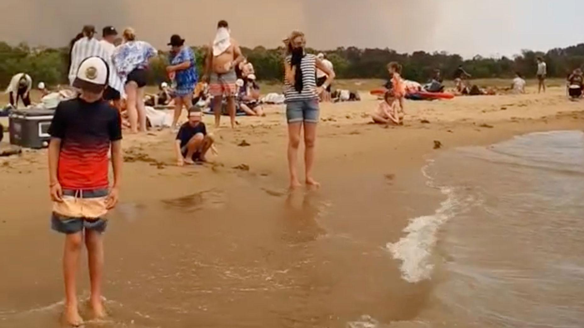 Several people are shown standing on a beach with many holding cloth to their faces to protect from the smoke.
