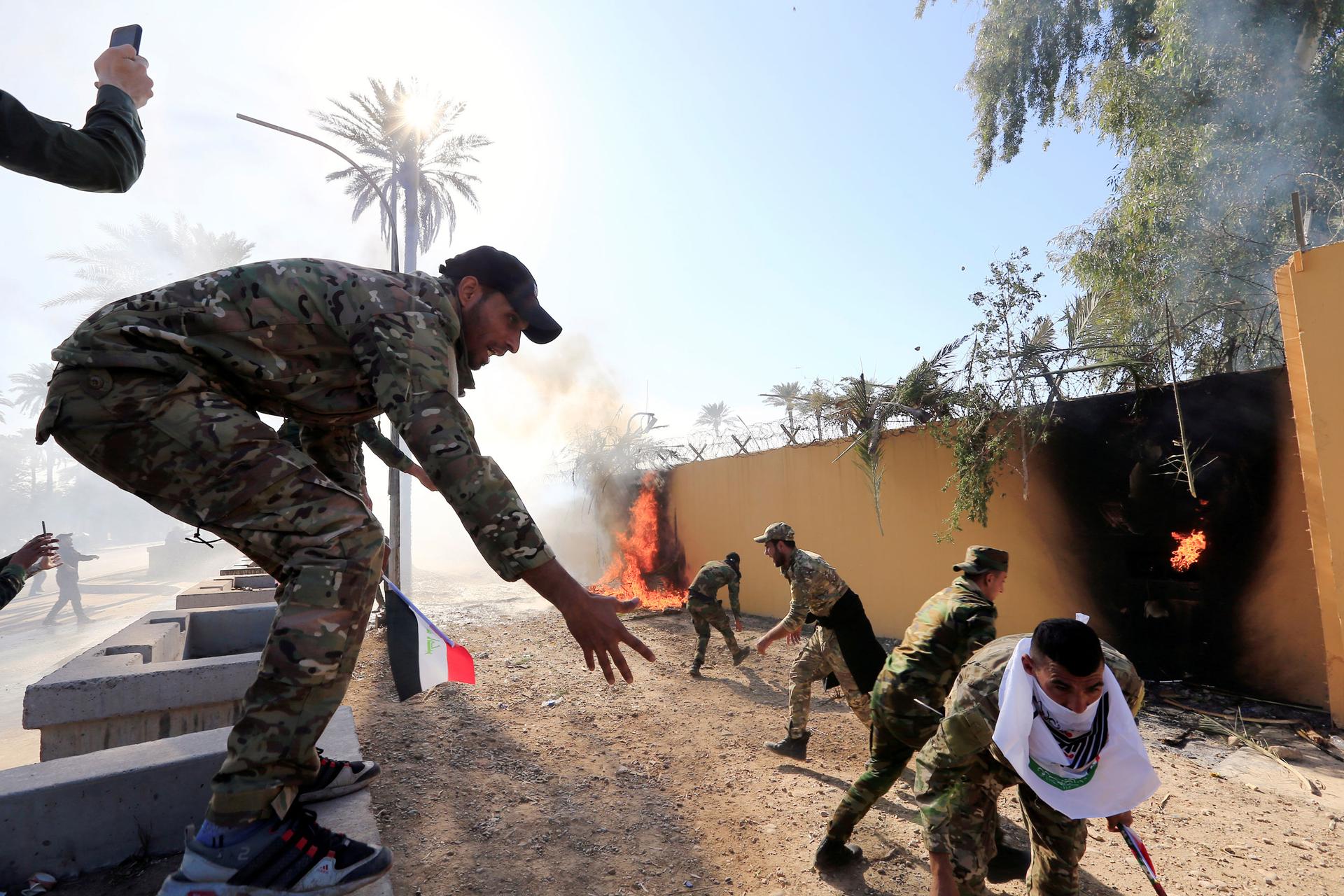 Several men are shown wearing military fatigues outside of a khaki-colored wall that has been set on fire.