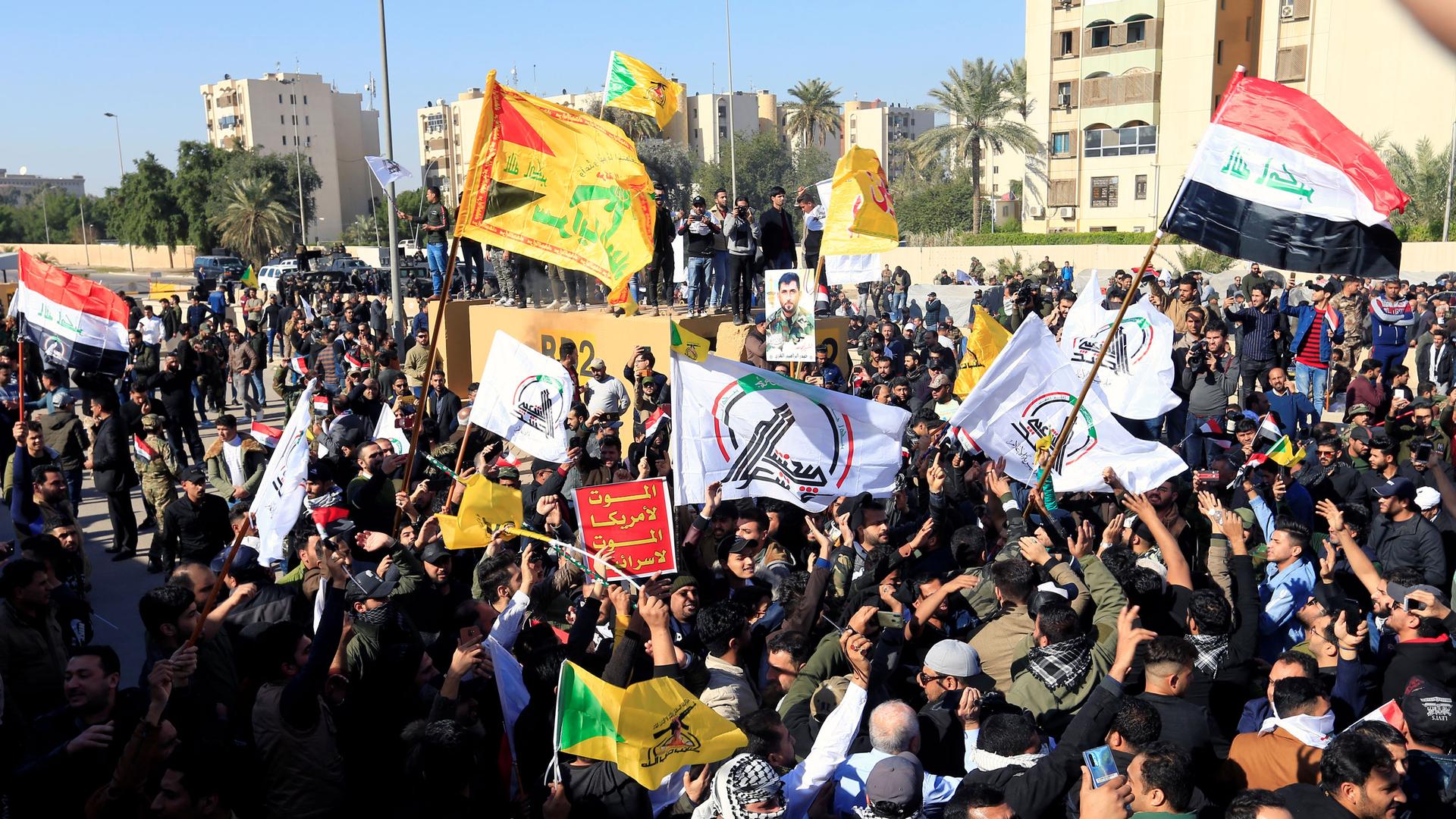 A large group of protesters are shown carrying several different flags outside of the khaki-colored walls of US Embassy in Iraq.