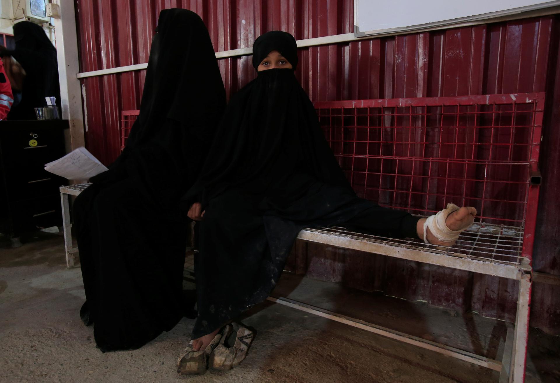 Two girls wearing black clothing sit on a bench in a clinic, one has a wounded foot wrapped in bandage