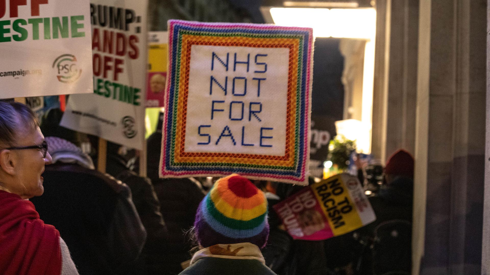 A protester stands with a sign reading "NHS NOT FOR SALE"