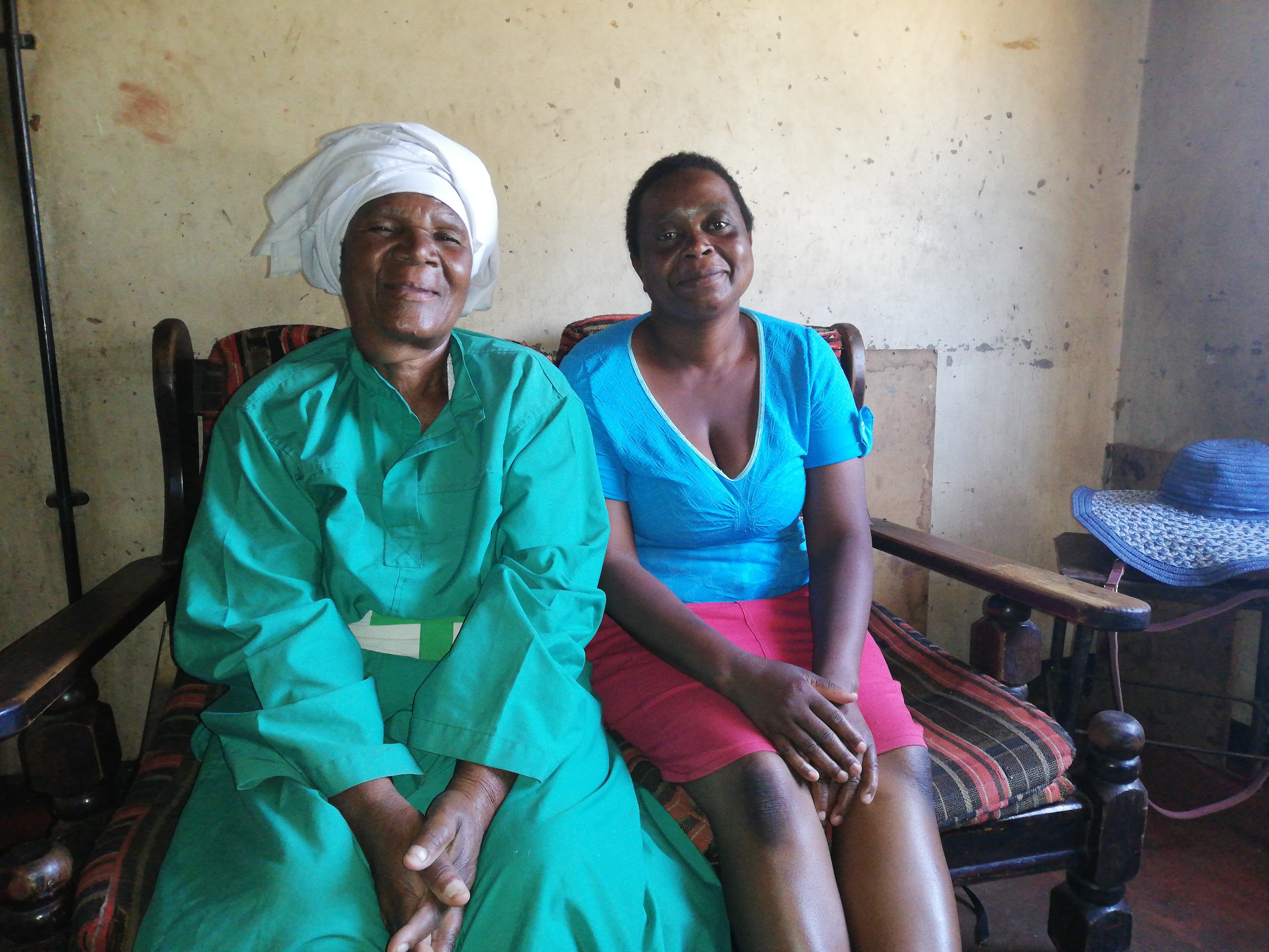 A woman wearing a green dress sits next to a woman wearing a blue shirt and pink skirt sit on a bench inside a clinic.