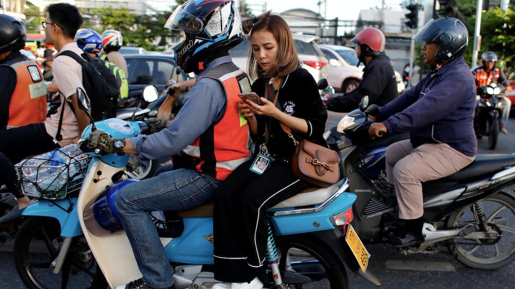 A woman checks her phone while riding on the back of a motorbike. 