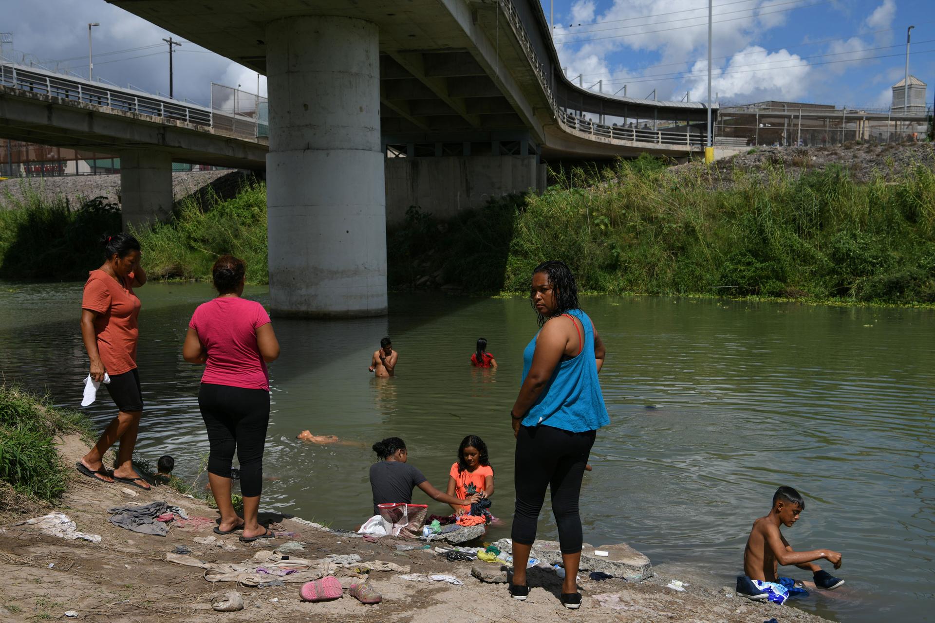 Several people are shown standing on the shore and in the Rio Grande river under a large bridge.