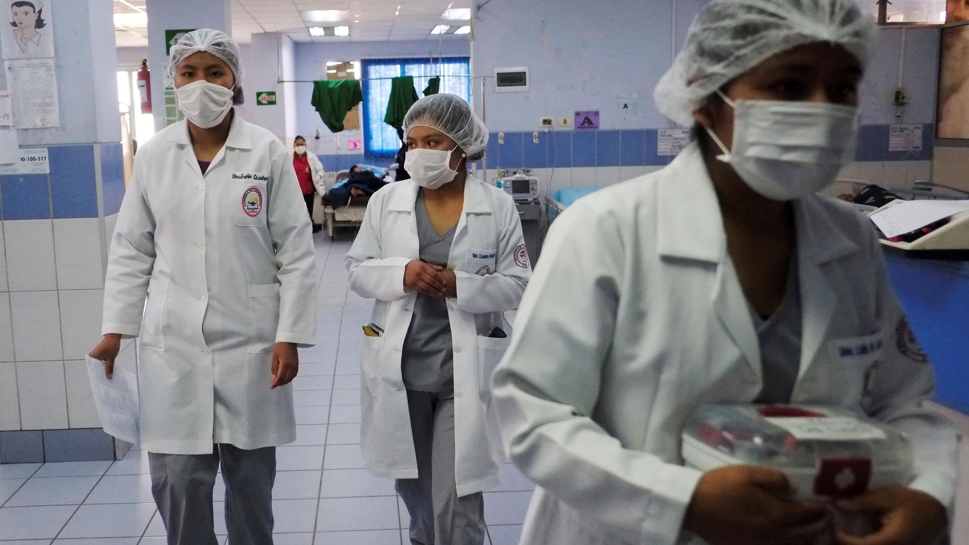 Three nurses are shown wearing white coats and hair coverings and walking in a hospital hallway.