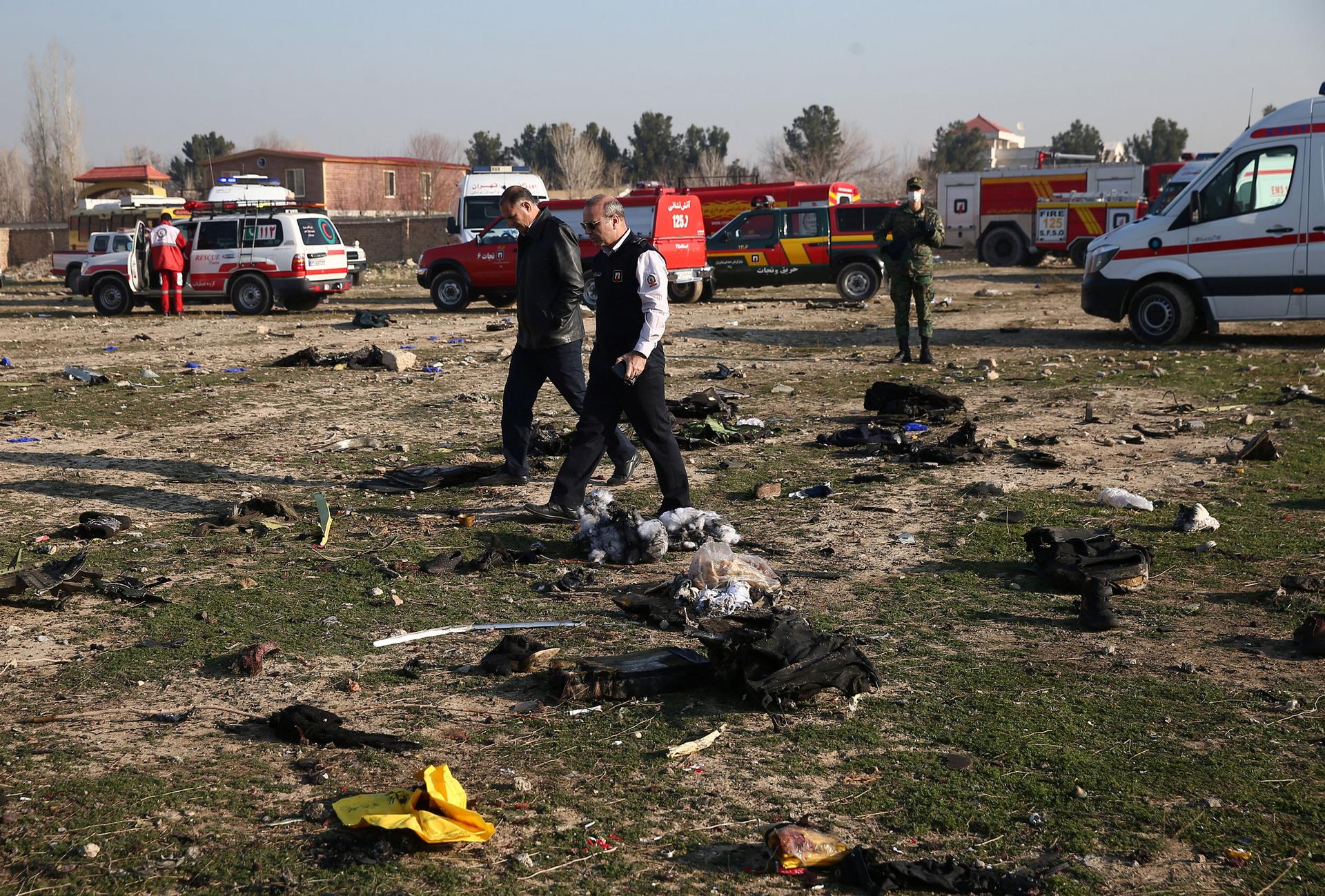 Two people are shown walking across a field strewn with reminant of a plane crash with several emergency vehicles in the background.