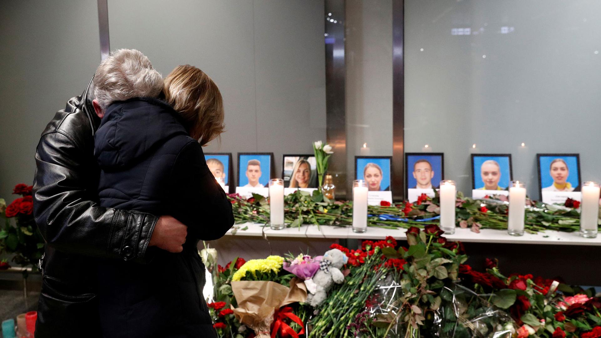 Two people are shown hugging while standing in front of a makeshift memorial for Ukraine Airlines flight that crashed.
