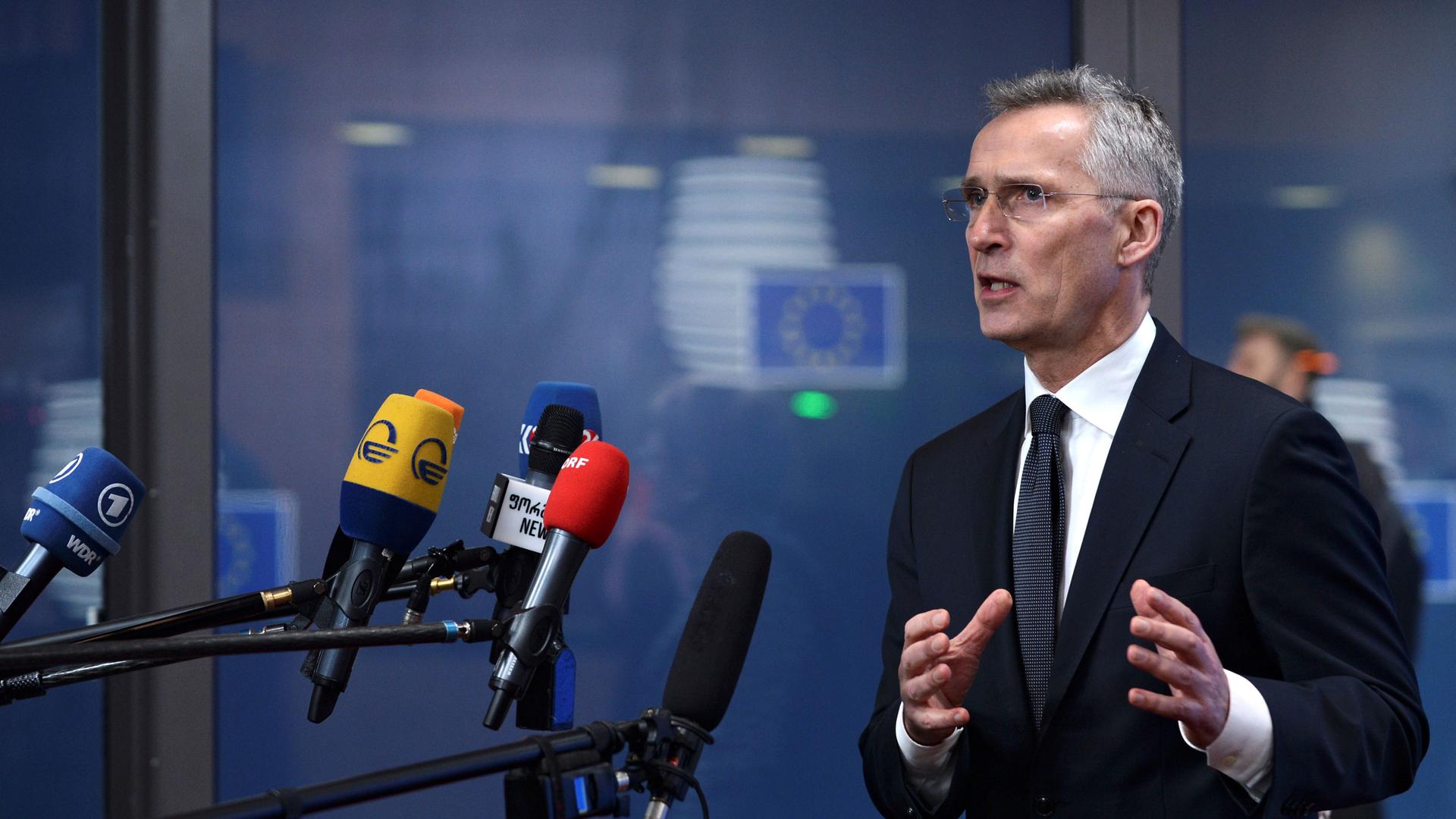 NATO Secretary General Jens Stoltenberg is shown wearing a dark suit and standing in front of several microphones.