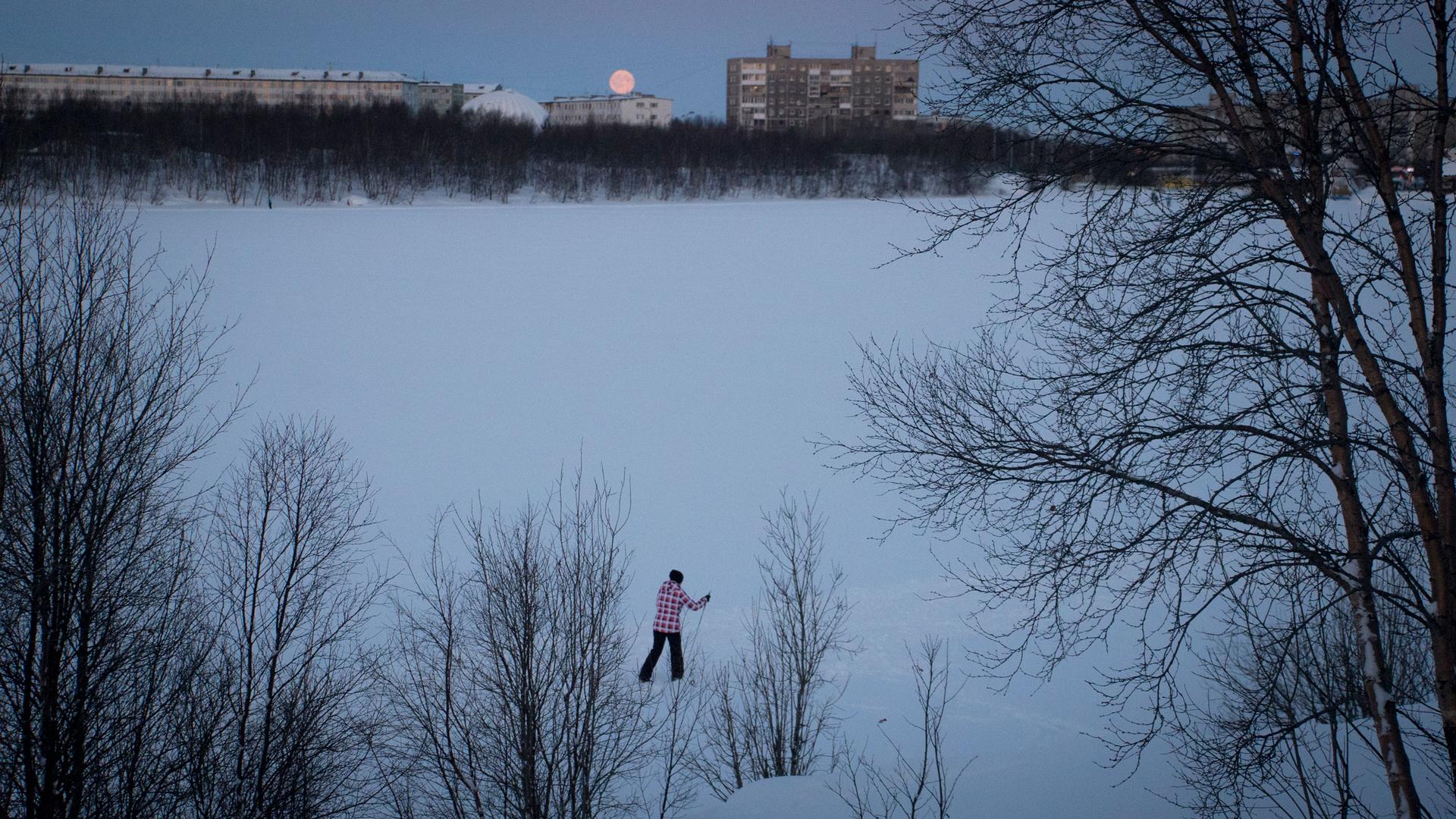 A person skis across a large snow-covered area with trees in the nearground and brick building and the moon in the background.