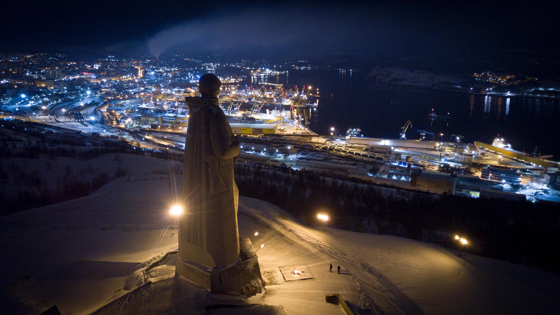 The Alyosha monument stands looking out over Murmansk off in the distance on a dark night.
