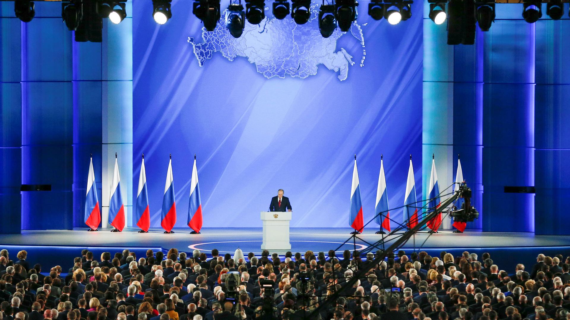 Russian President Vladimir Putin is shown standing on a large stage with several Russian flags behind him.