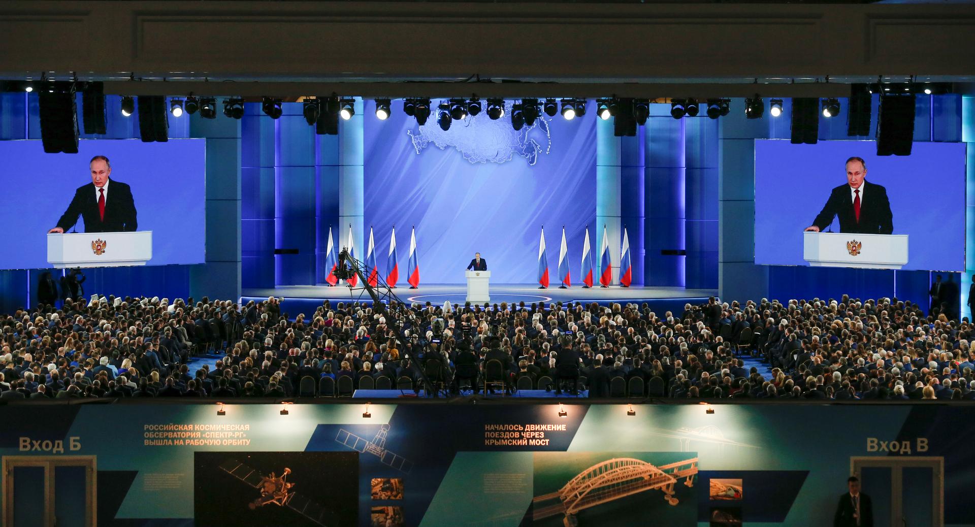 A man speaks from a podium in front of an audience. He is flanked by Russian flags and screens