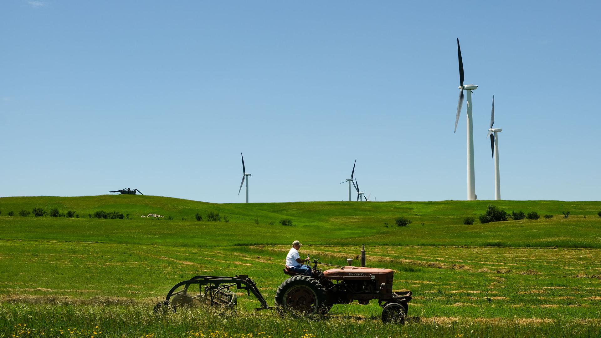 A farm tractor is shown in a large green field with wind turbines in the background.